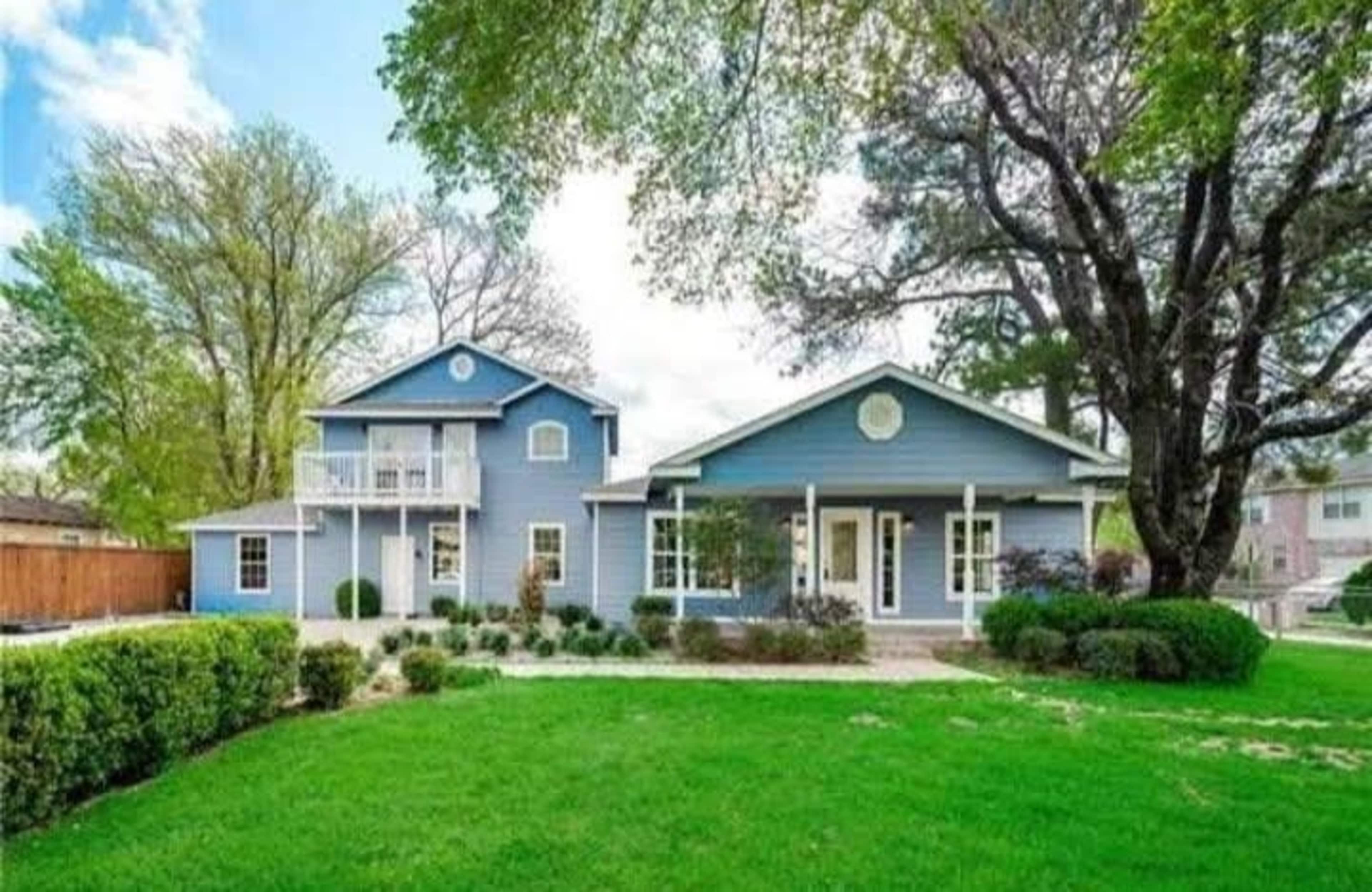 The image shows a large blue house with a two-story section on the left and a single-story section on the right, set in a landscaped yard with green grass and trees.