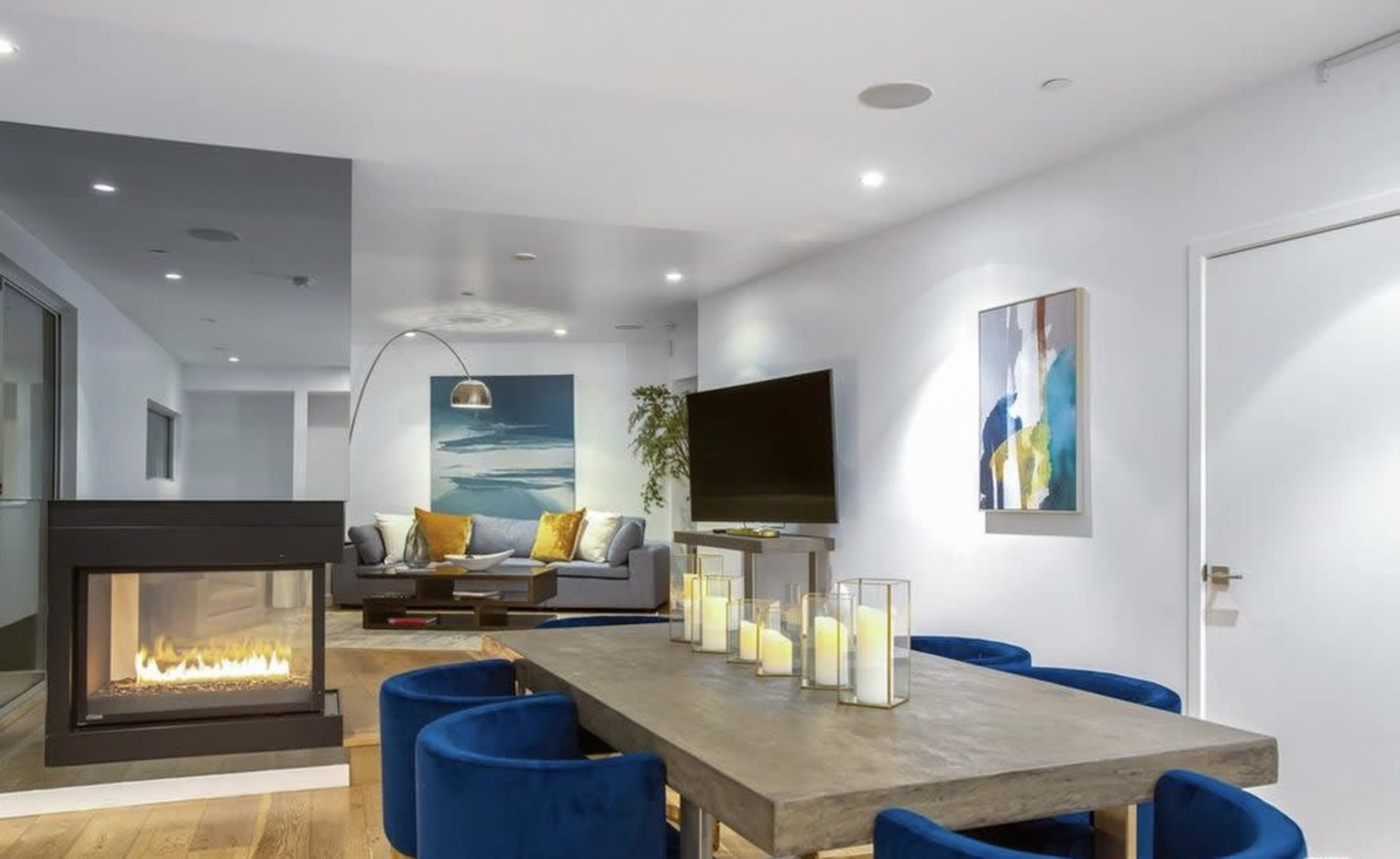 The image shows a modern living space featuring a fireplace, a dining table surrounded by blue velvet chairs, and a lounge area with a couch and artwork on the walls.