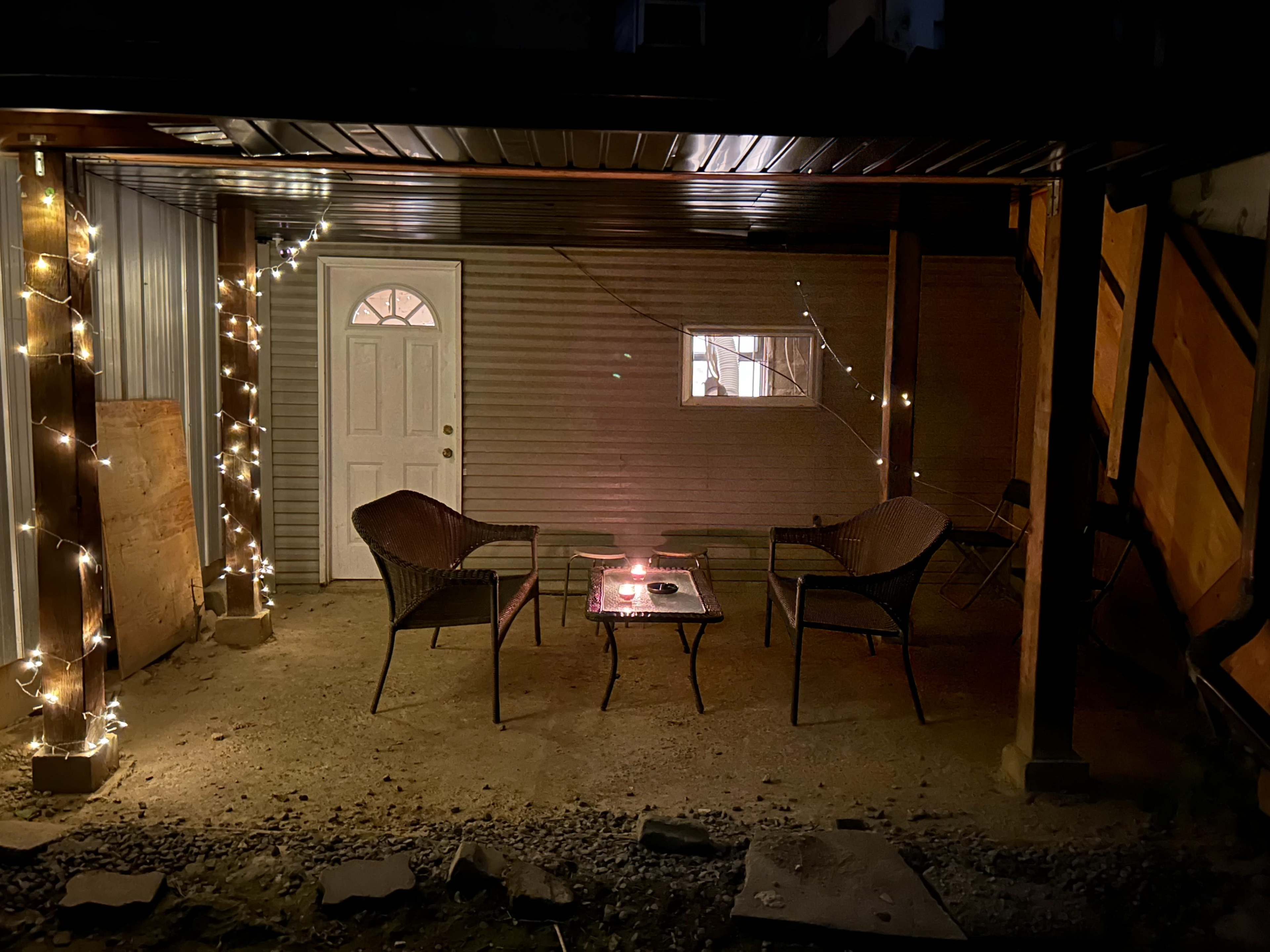 A covered outdoor area features two wicker chairs facing a glass coffee table, adorned with string lights and a door leading to an interior space.