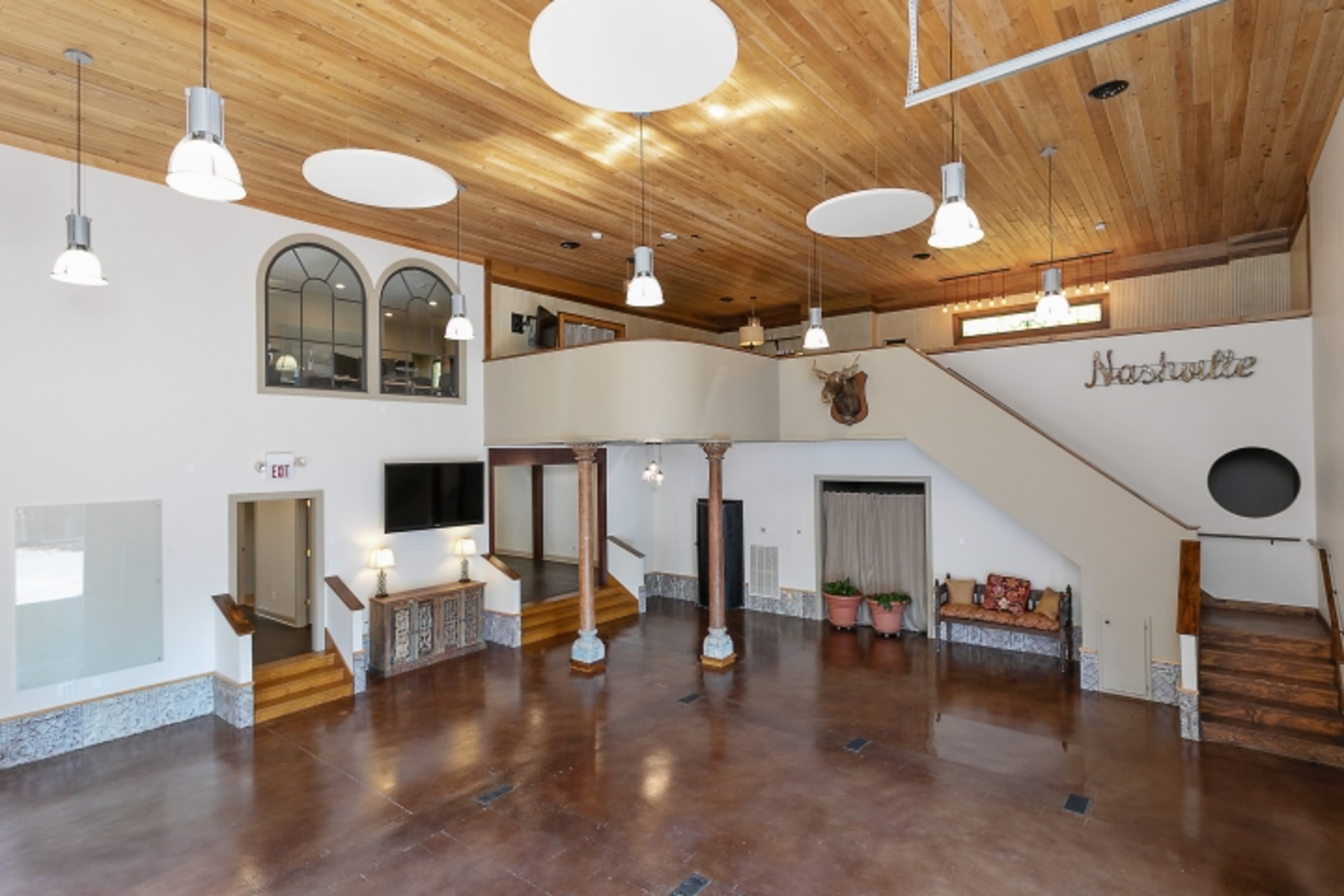 The image shows an open, spacious interior with a wooden ceiling, large windows, and a staircase leading to a second level.