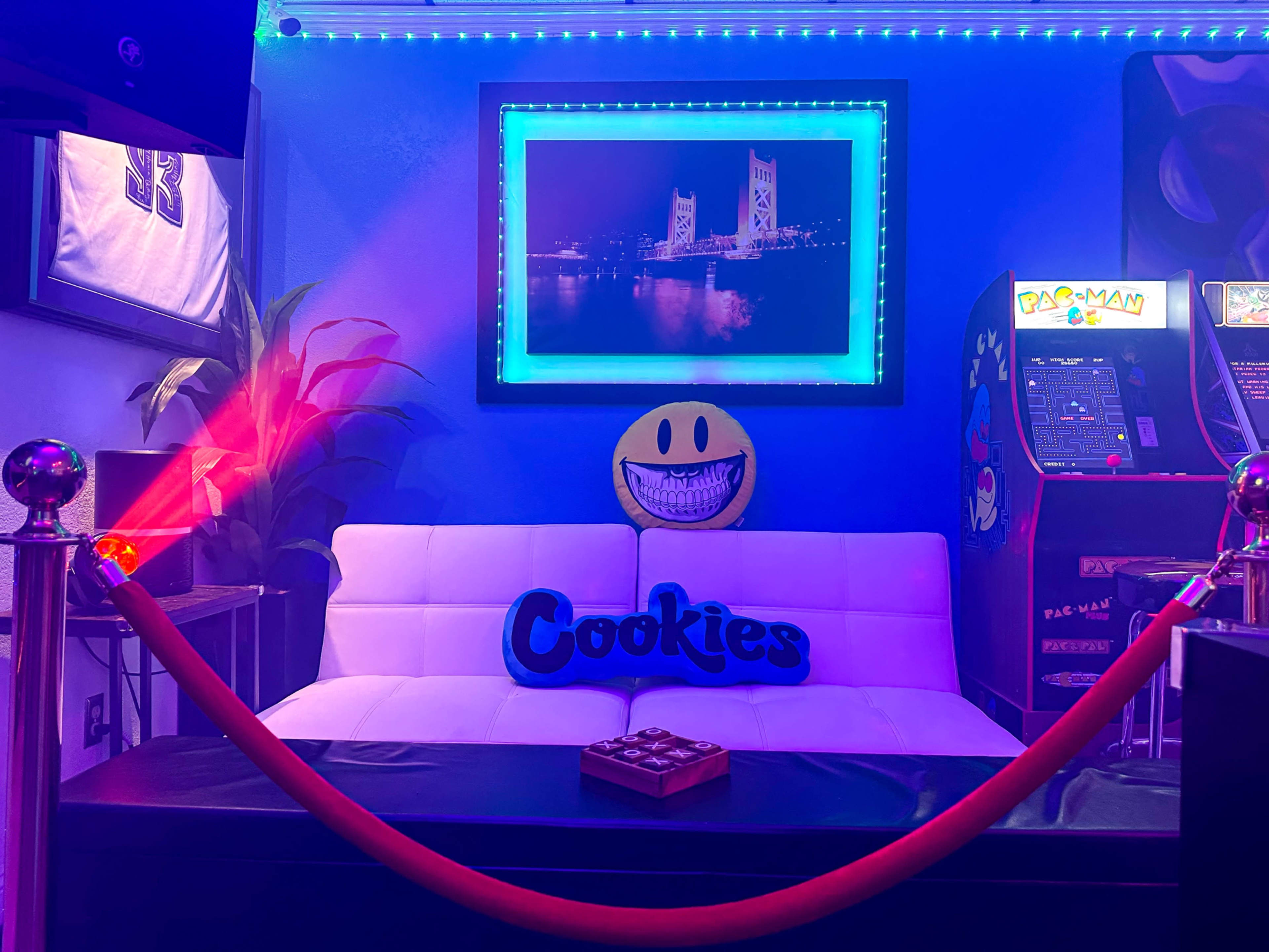 The image shows a lounge area with a white couch, a smiling emoji pillow, a "Cookies" sign, arcade machines, and colored LED lighting.