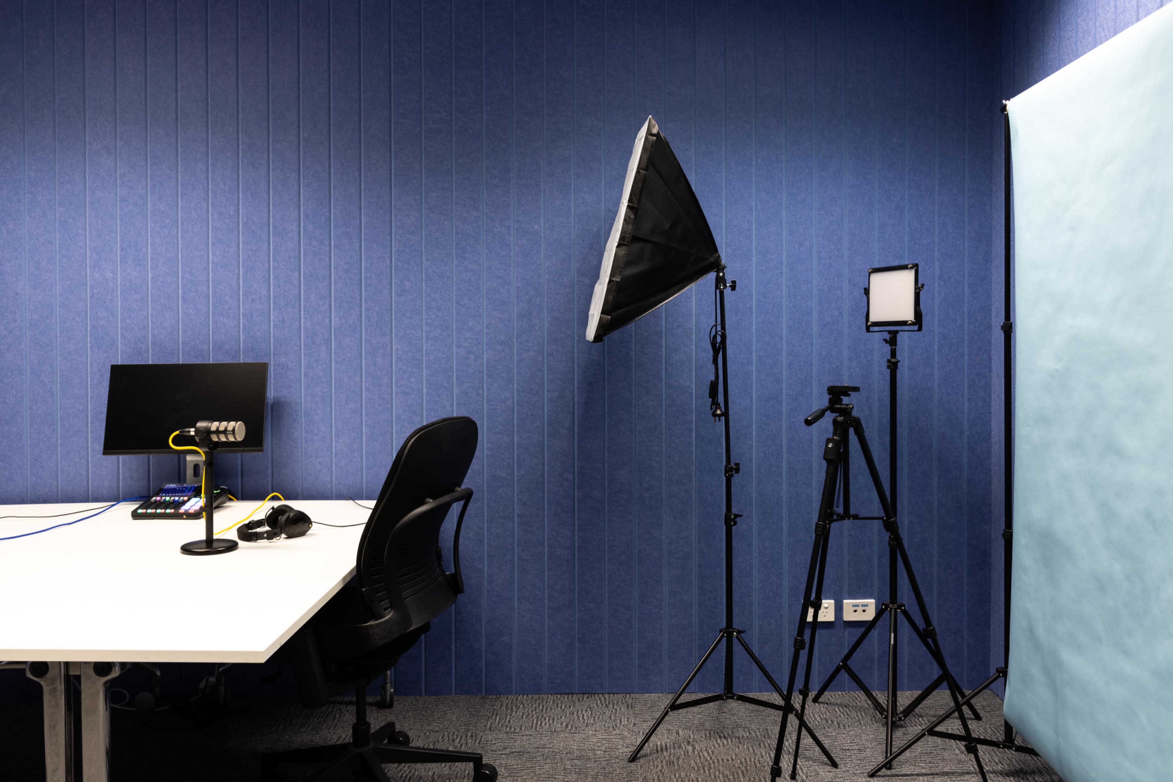 The image shows a small recording studio with a desk equipped with a computer, a microphone, and a chair, alongside lighting equipment and a backdrop.