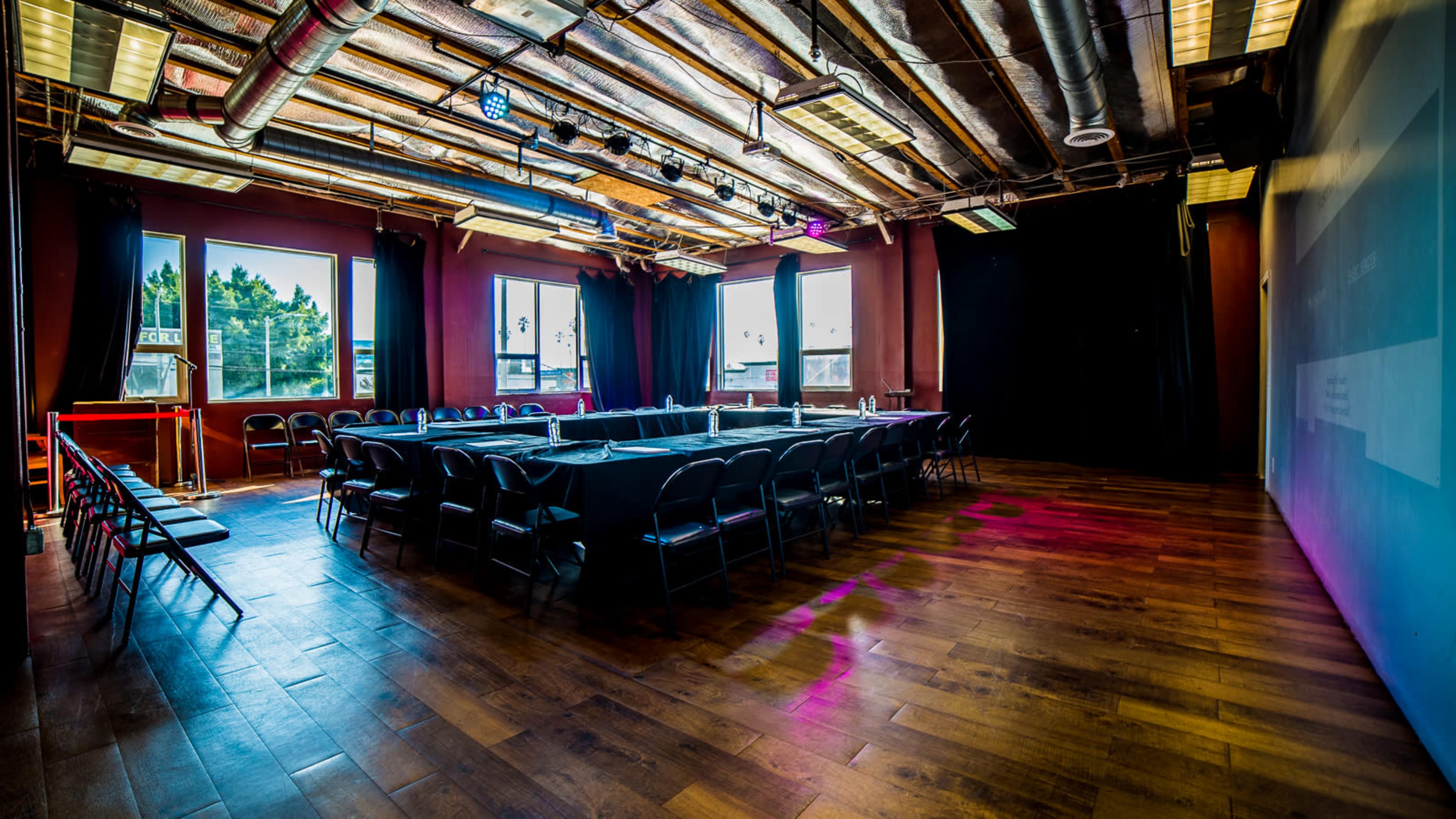 A conference room is set up with a large rectangular table surrounded by black chairs under a wooden ceiling and bright windows.