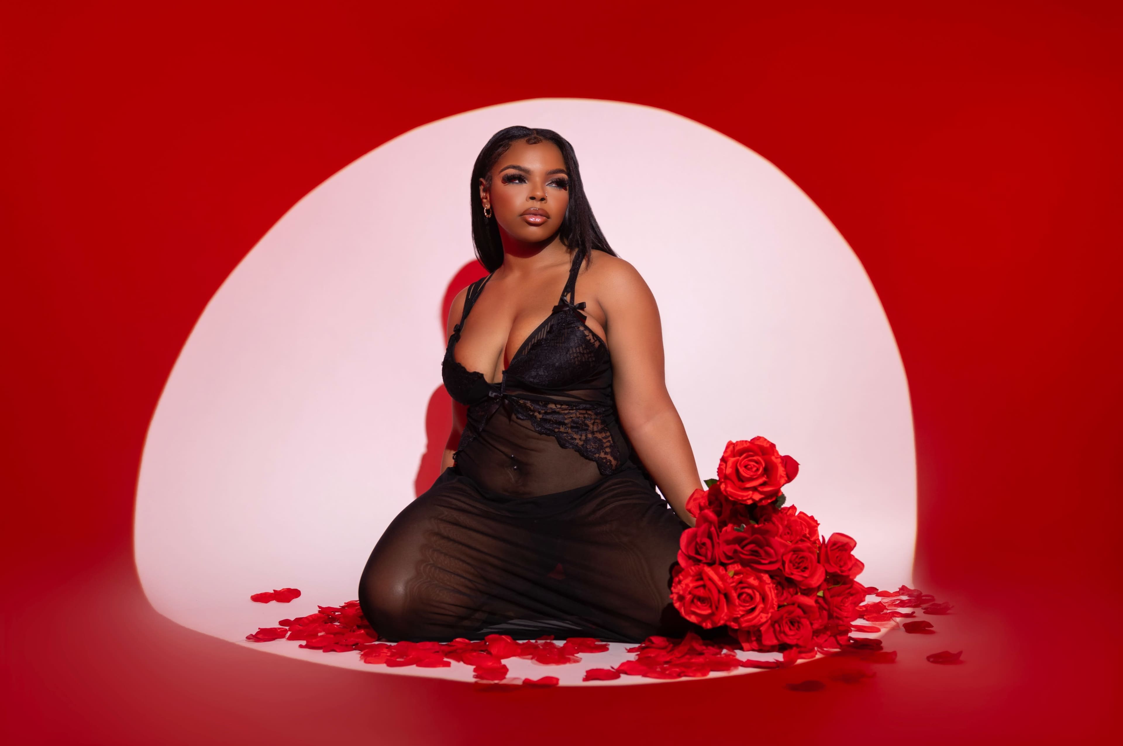 A woman in a black sheer dress sits on the floor surrounded by rose petals, holding a large bouquet of red roses against a vibrant red backdrop.