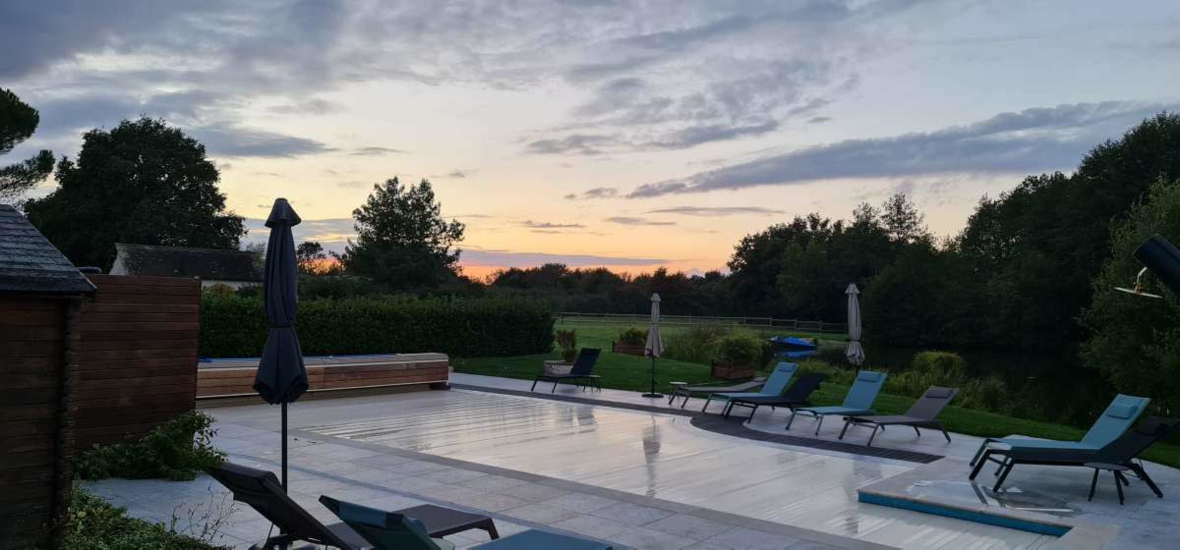 The scene depicts a tranquil outdoor area featuring a swimming pool surrounded by lounge chairs, with a sunset casting soft light over the landscape.