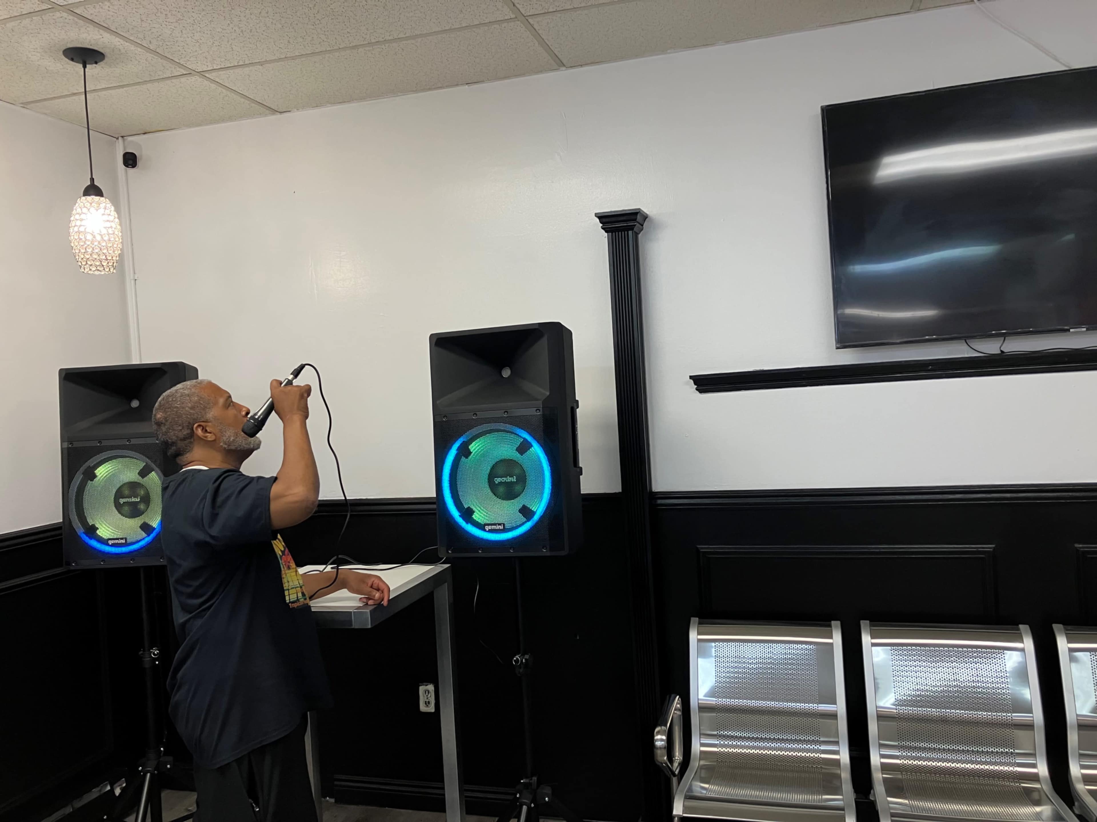 A man stands in front of two speakers, holding a microphone, and looking up at a television on the wall.