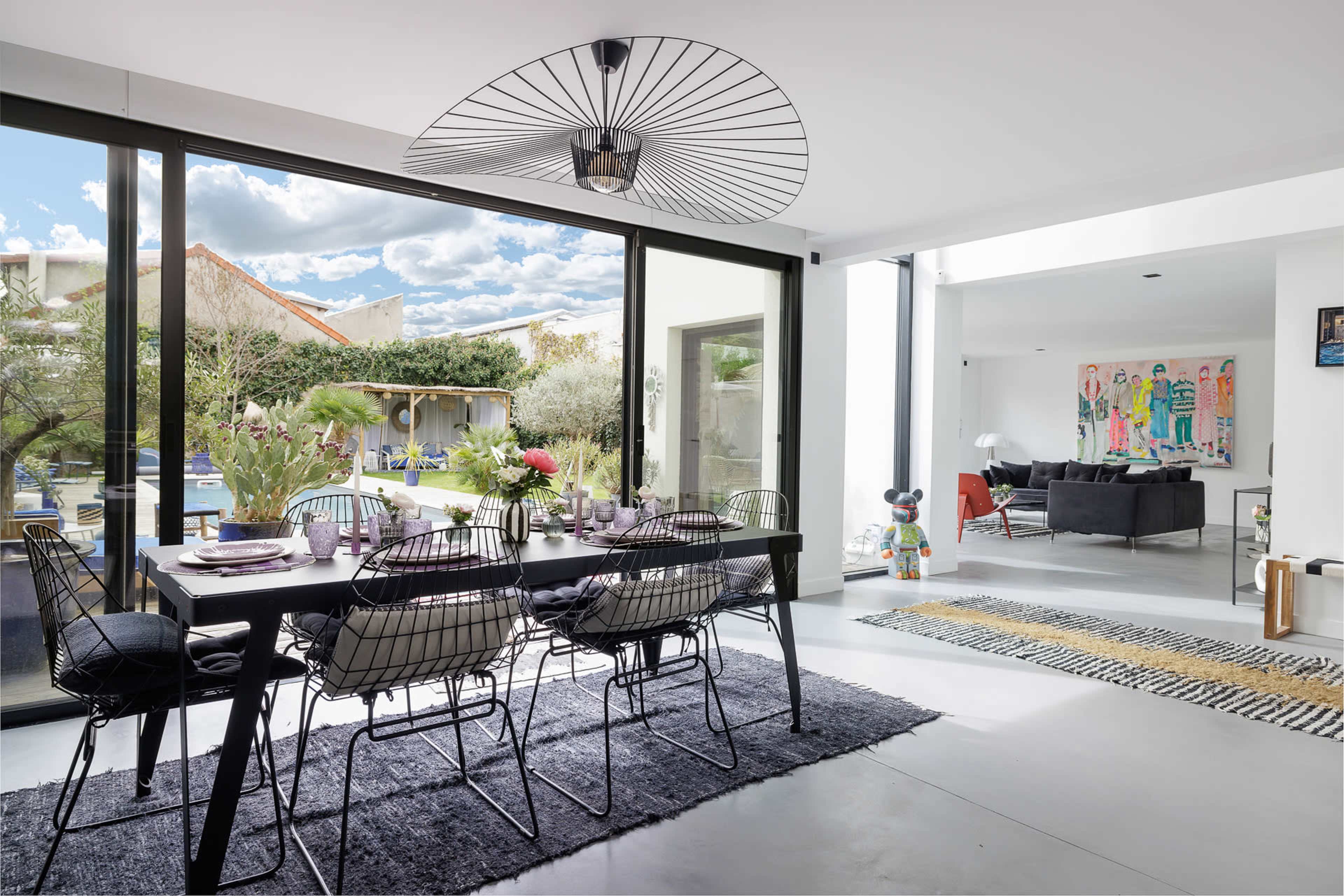 A modern dining area features a black table surrounded by wire chairs, with large windows overlooking a garden and pool.