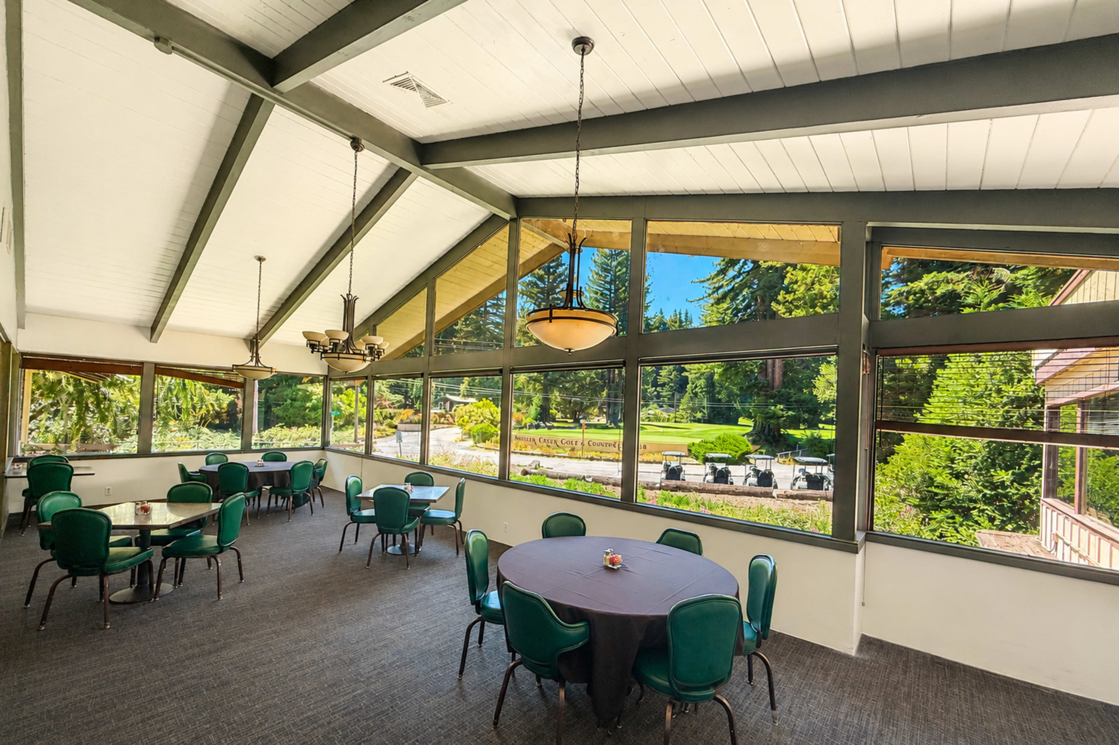 The image shows a spacious indoor dining area with round tables and green chairs, featuring large windows that overlook a landscaped outdoor space.