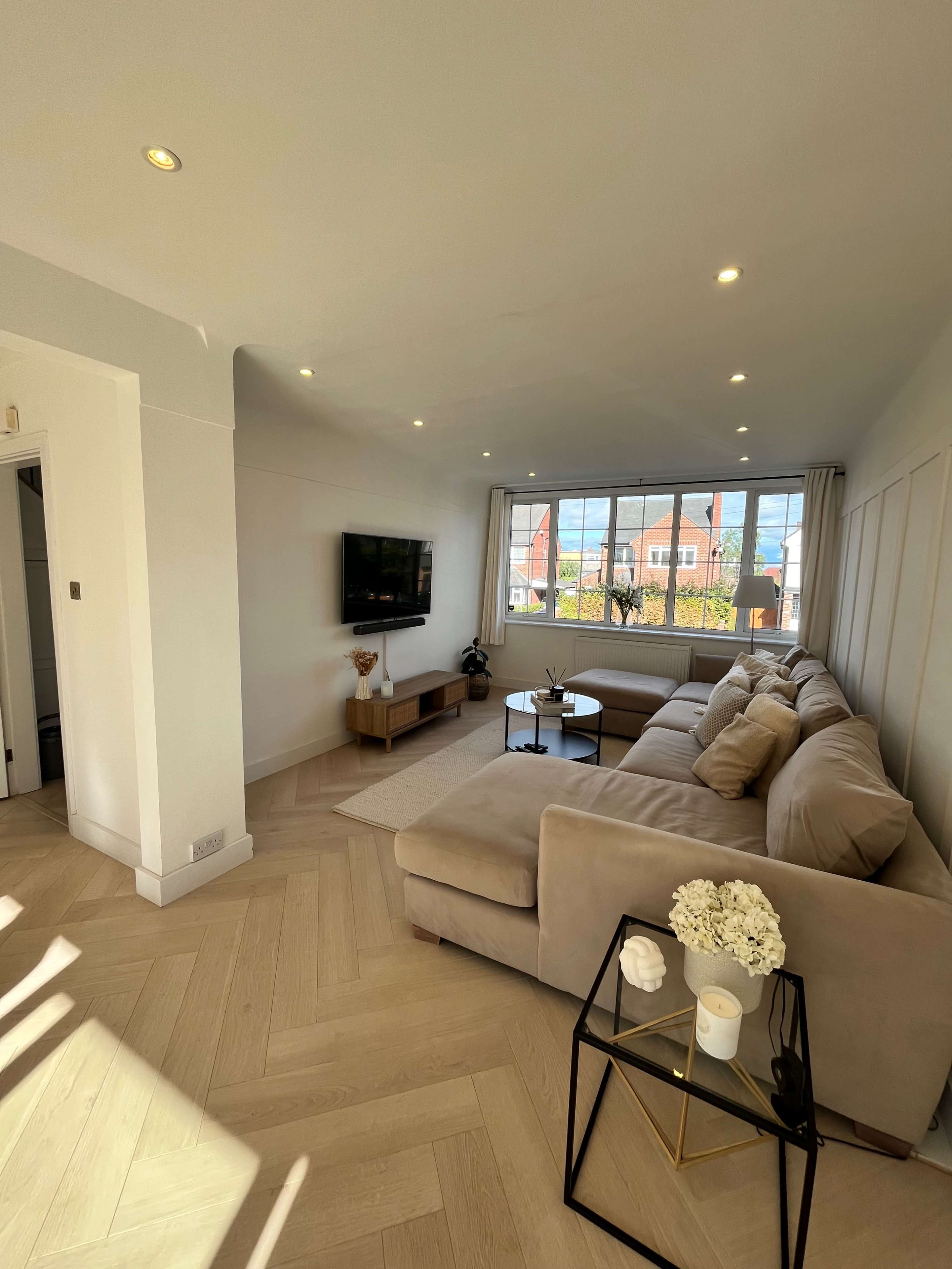 A modern living room features a light-colored sectional sofa, a coffee table, and a wall-mounted television, with large windows allowing natural light to fill the space.