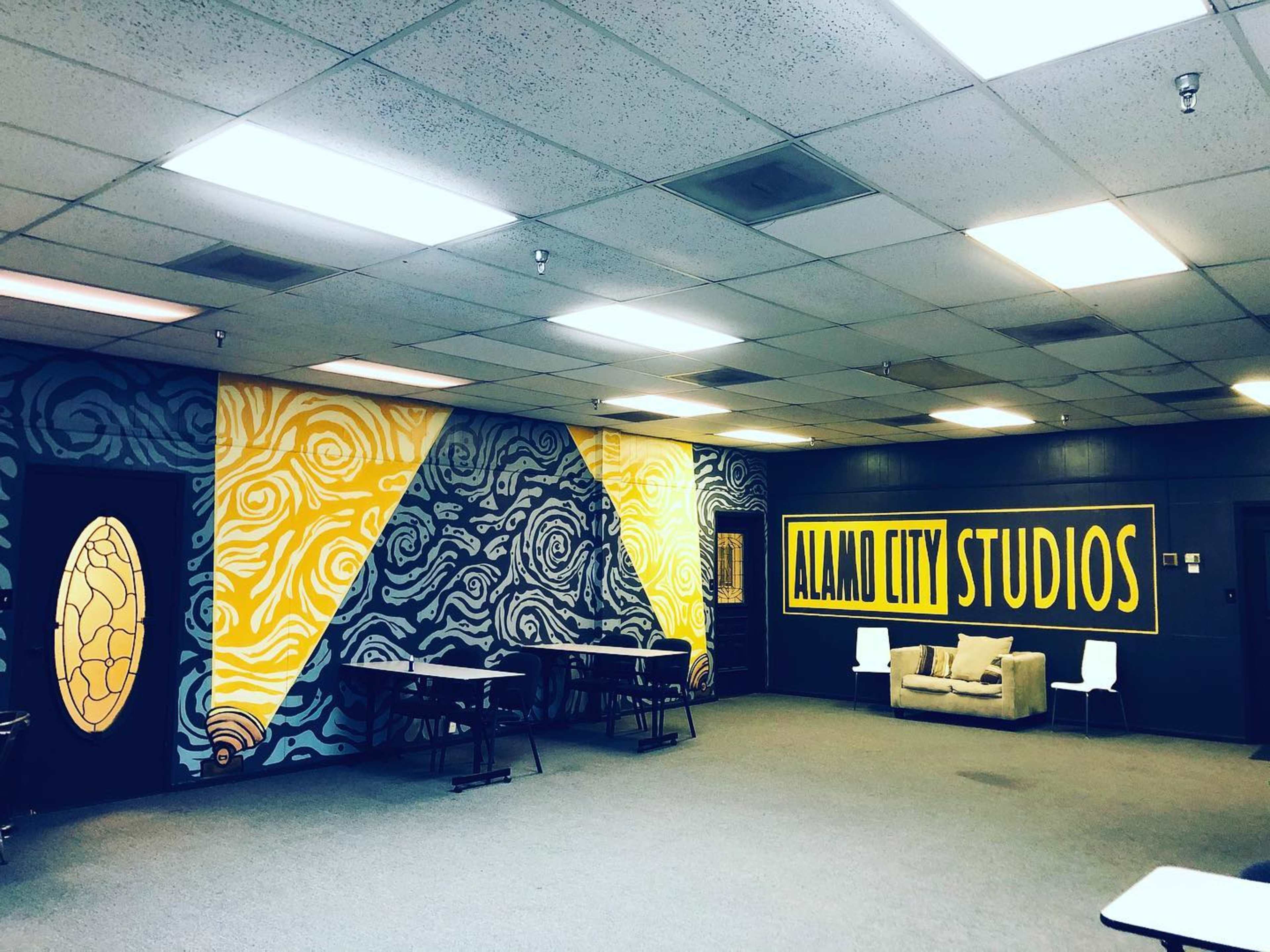 The image shows an interior space labeled "Alamo City Studios," featuring colorful murals on the walls and empty tables and chairs arranged in the area.