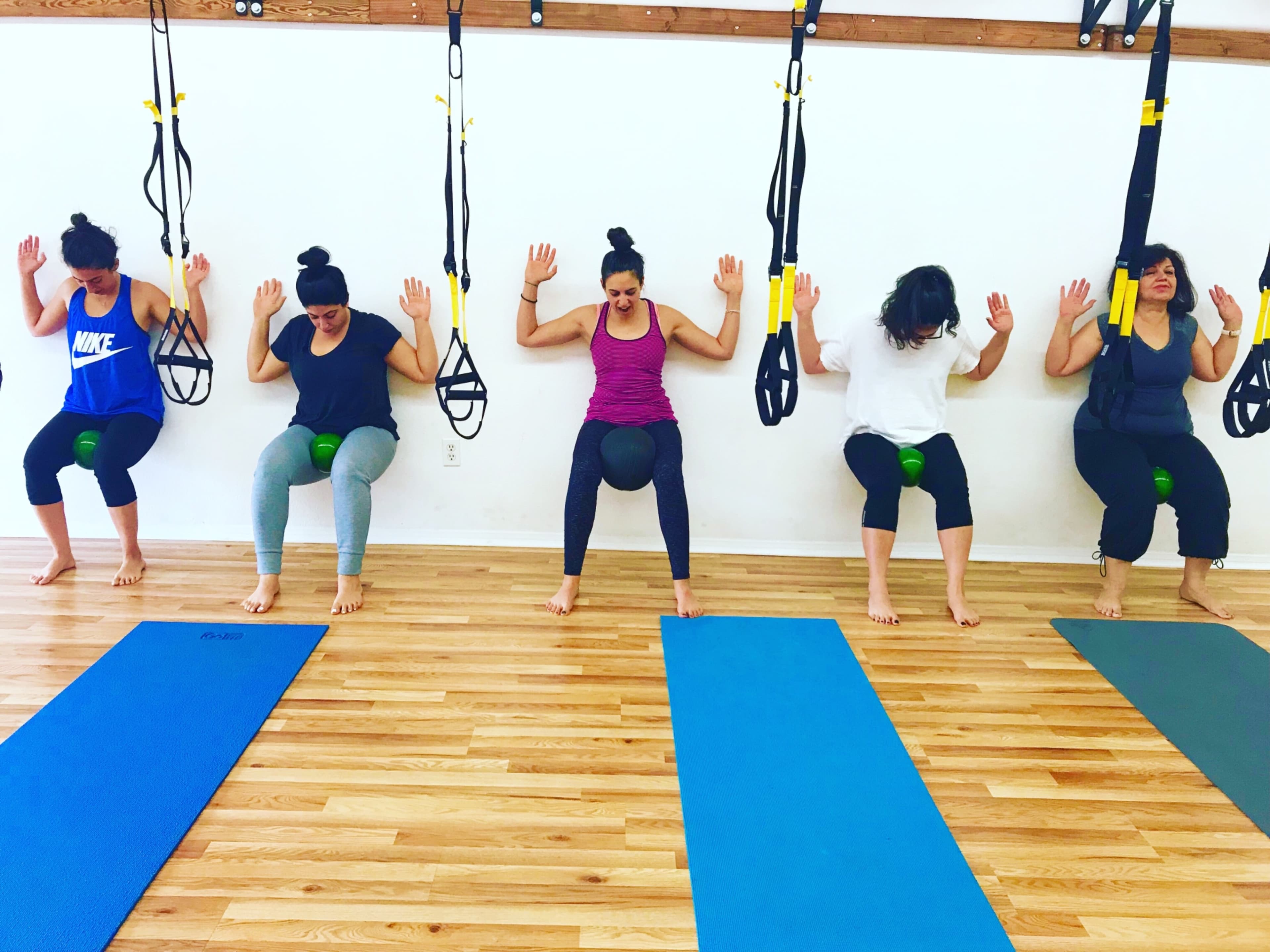 Five women are practicing exercises on stability balls while suspended with resistance bands in a fitness studio.
