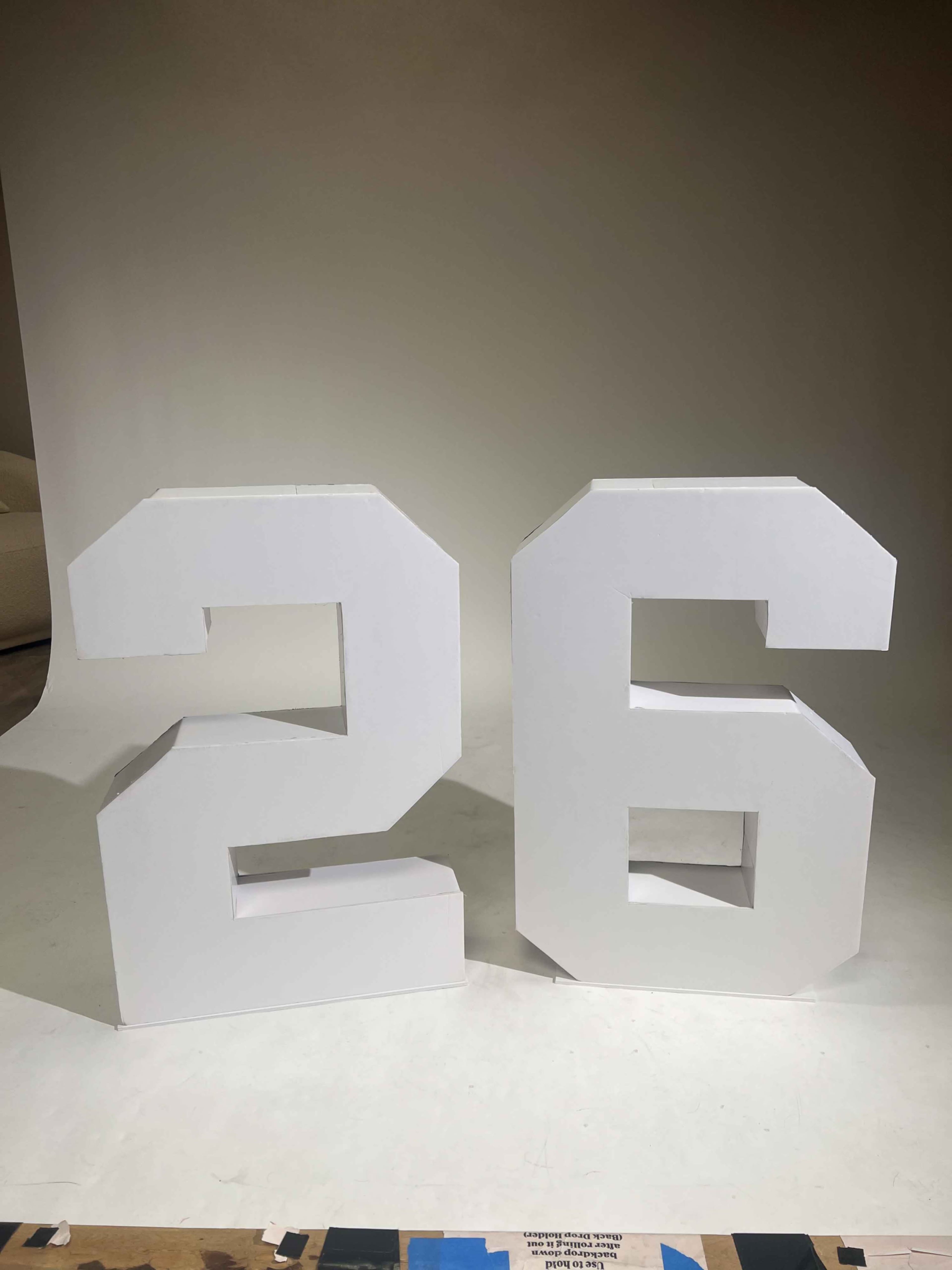 The image features large, three-dimensional white numbers "2" and "6" standing on a flat surface against a neutral background.