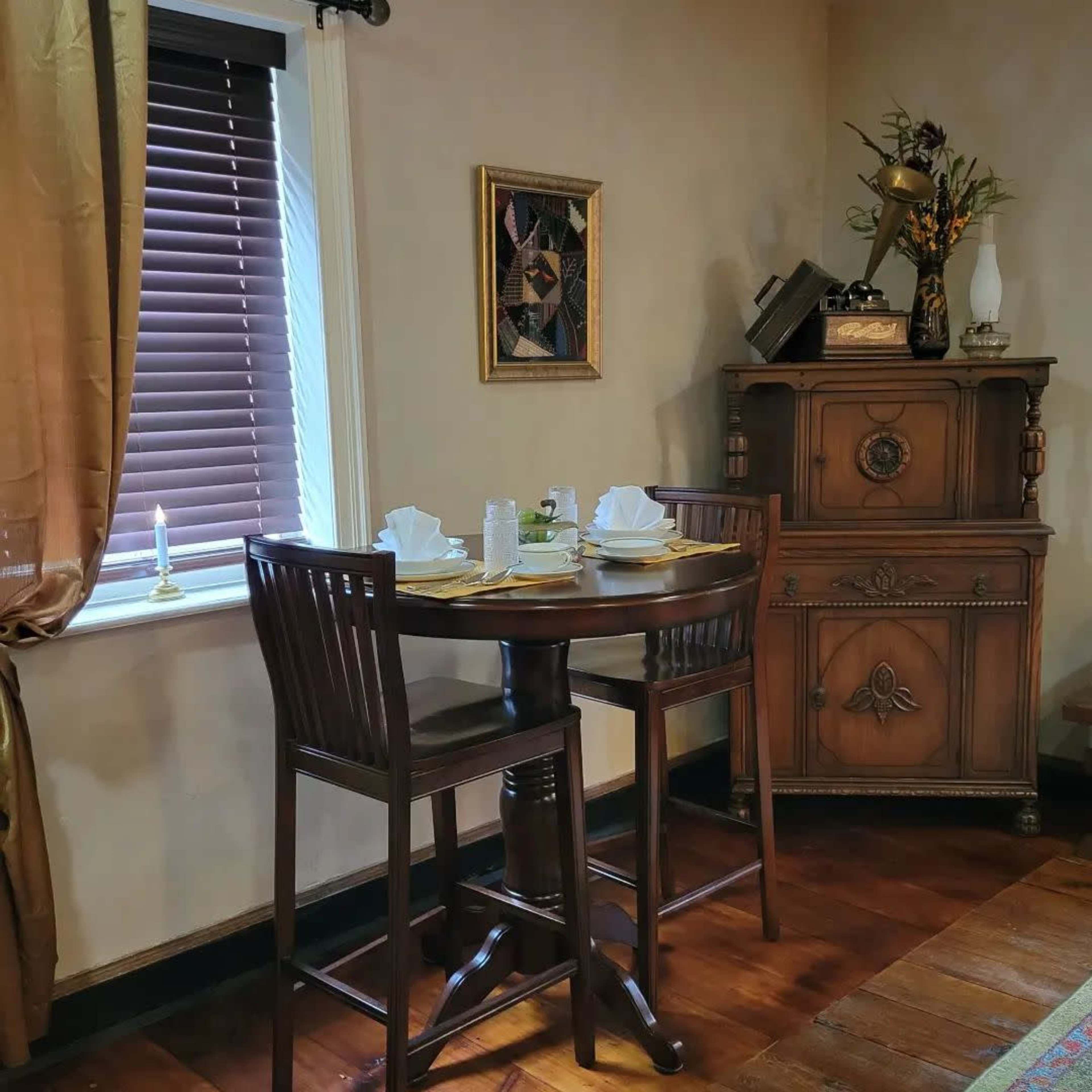 A wooden dining table with two chairs is set for a meal, accompanied by a decorative cabinet and window with blinds.