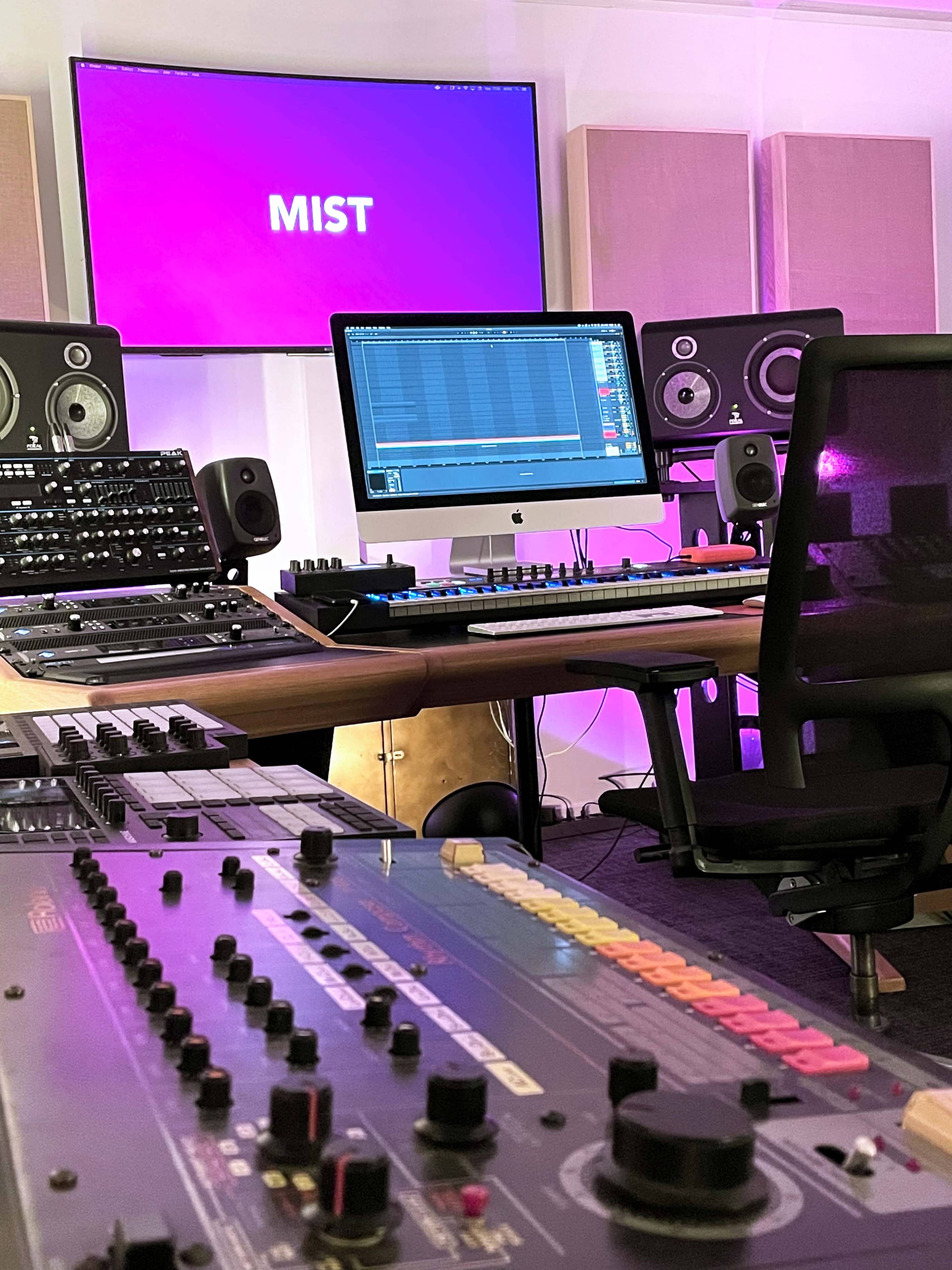 The image shows a music studio with various audio equipment, a computer monitor displaying the word "MIST," and a keyboard positioned in front of a chair.