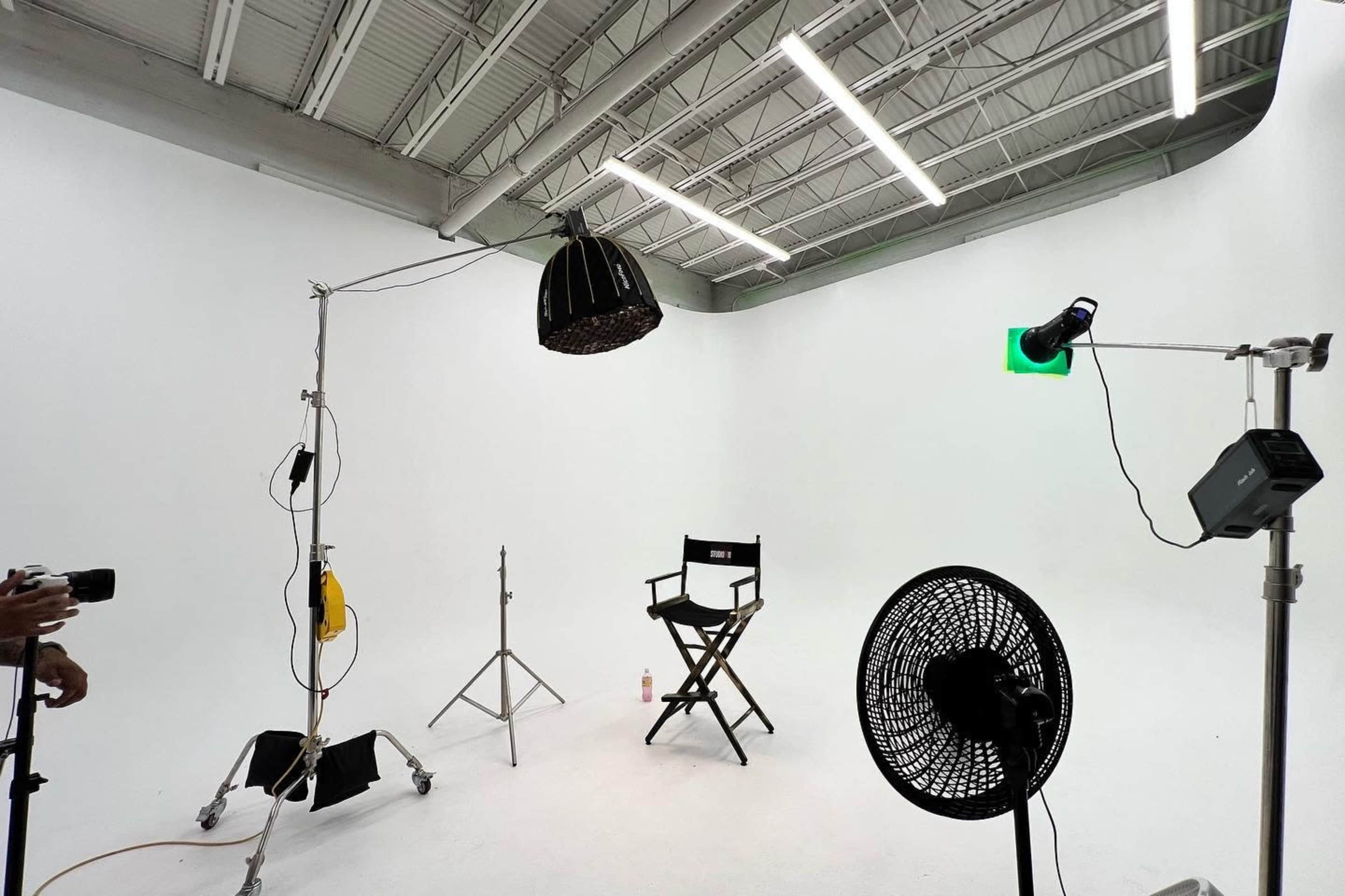 A minimalist photography studio setup with a director's chair, lighting equipment, and a cooling fan against a white background.