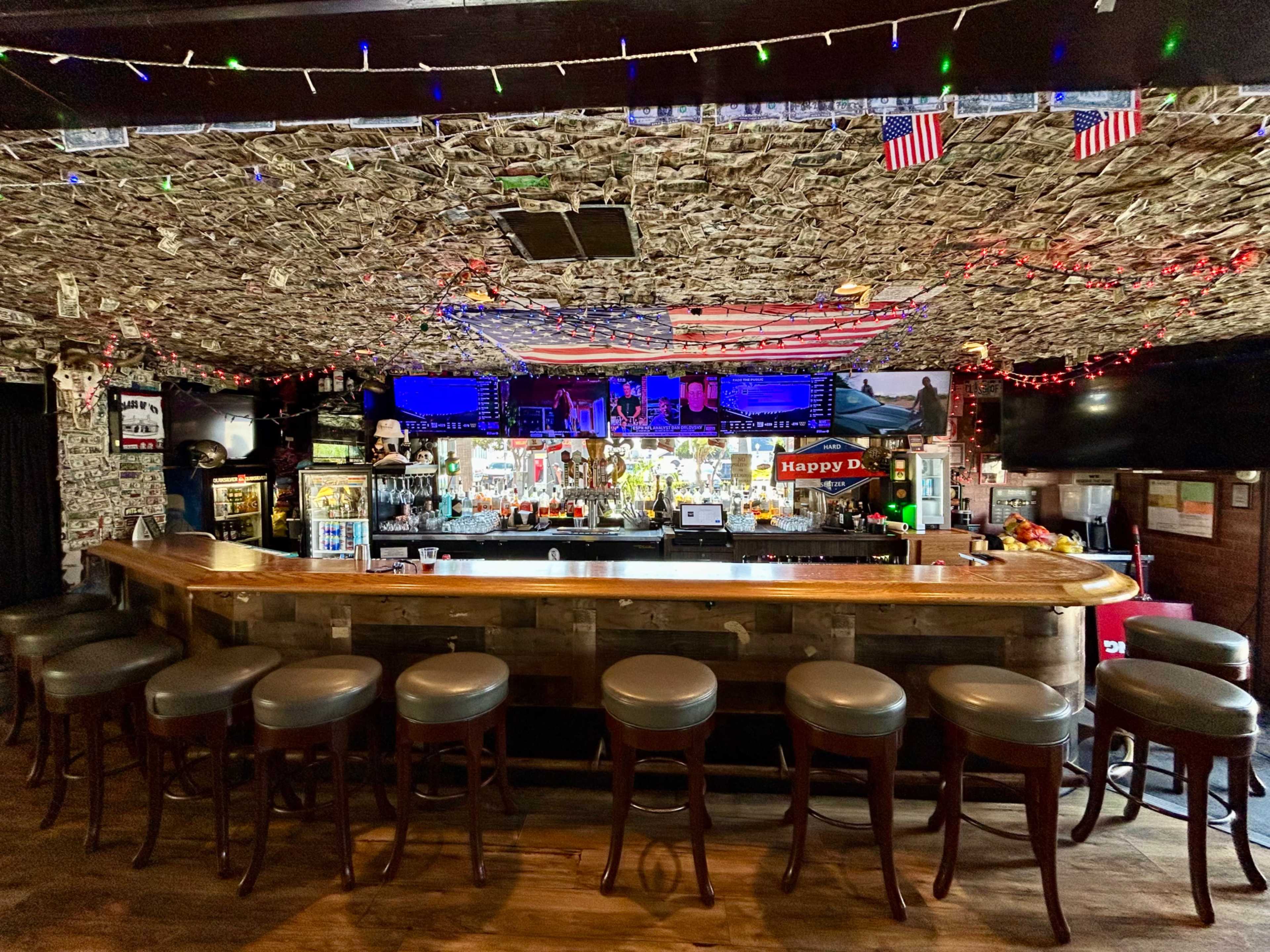 The image shows a bar with a wooden countertop and stools, decorated with dollar bills on the ceiling and a variety of colorful lights and flags hanging around the bar area.