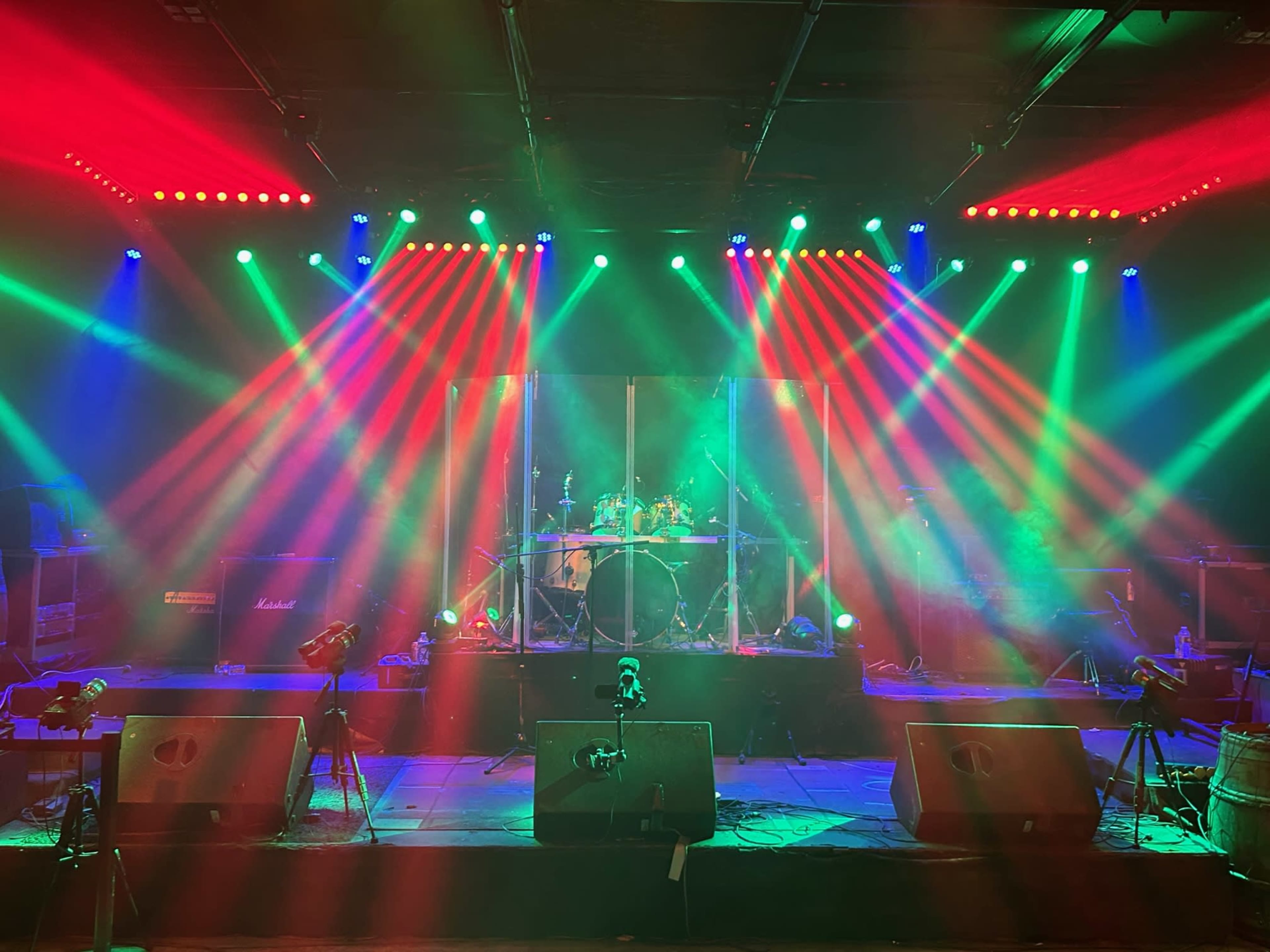 The stage is set with colorful lights shining down on a drum kit, surrounded by amplifiers and microphone stands.