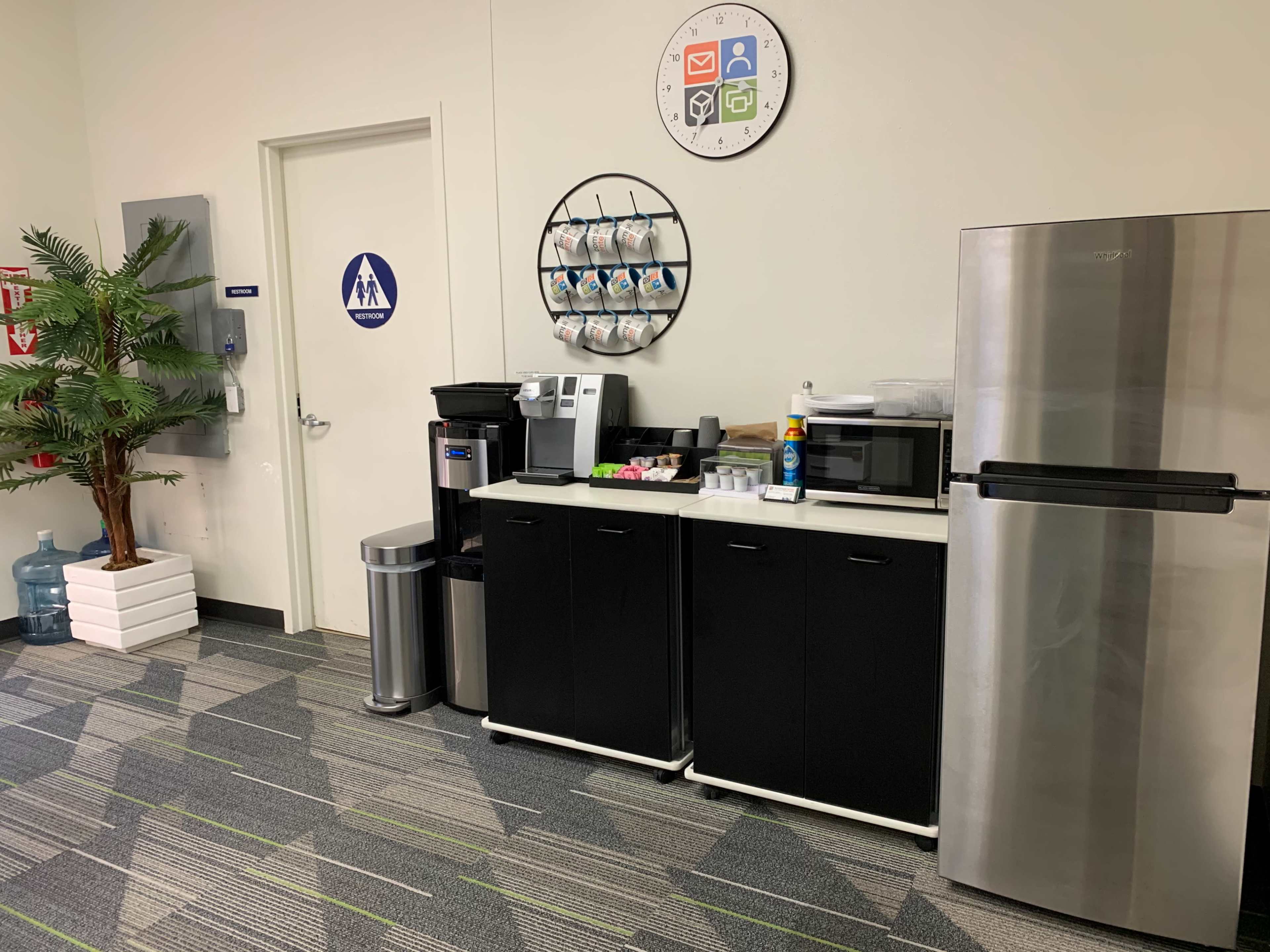 A break area with a coffee machine, microwave, refrigerator, and a wall-mounted organizer for cups, next to a restroom door.