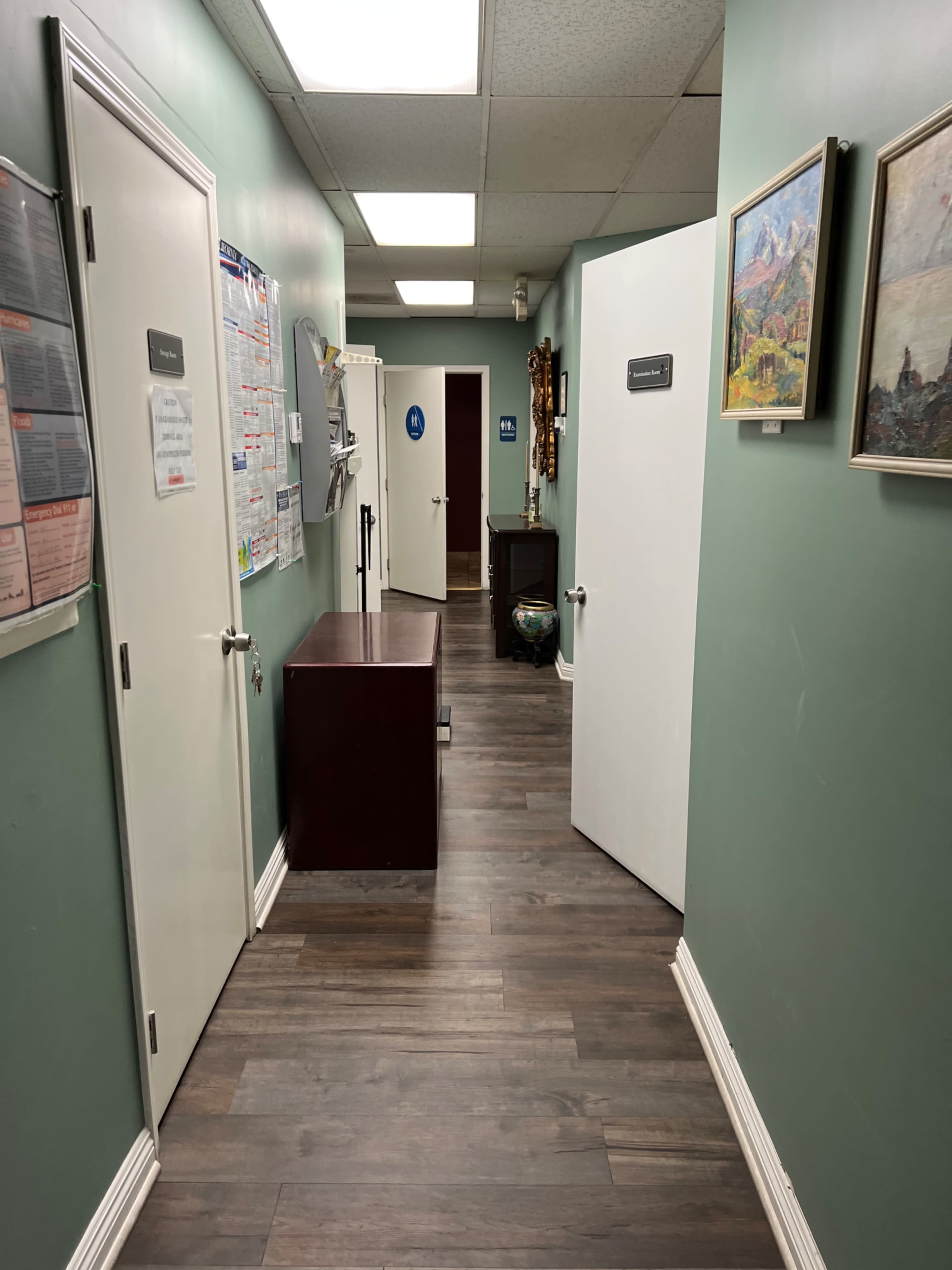 A hallway features doors on both sides, a wooden cabinet against the wall, and framed artwork hanging, with light-colored walls and a wooden floor.