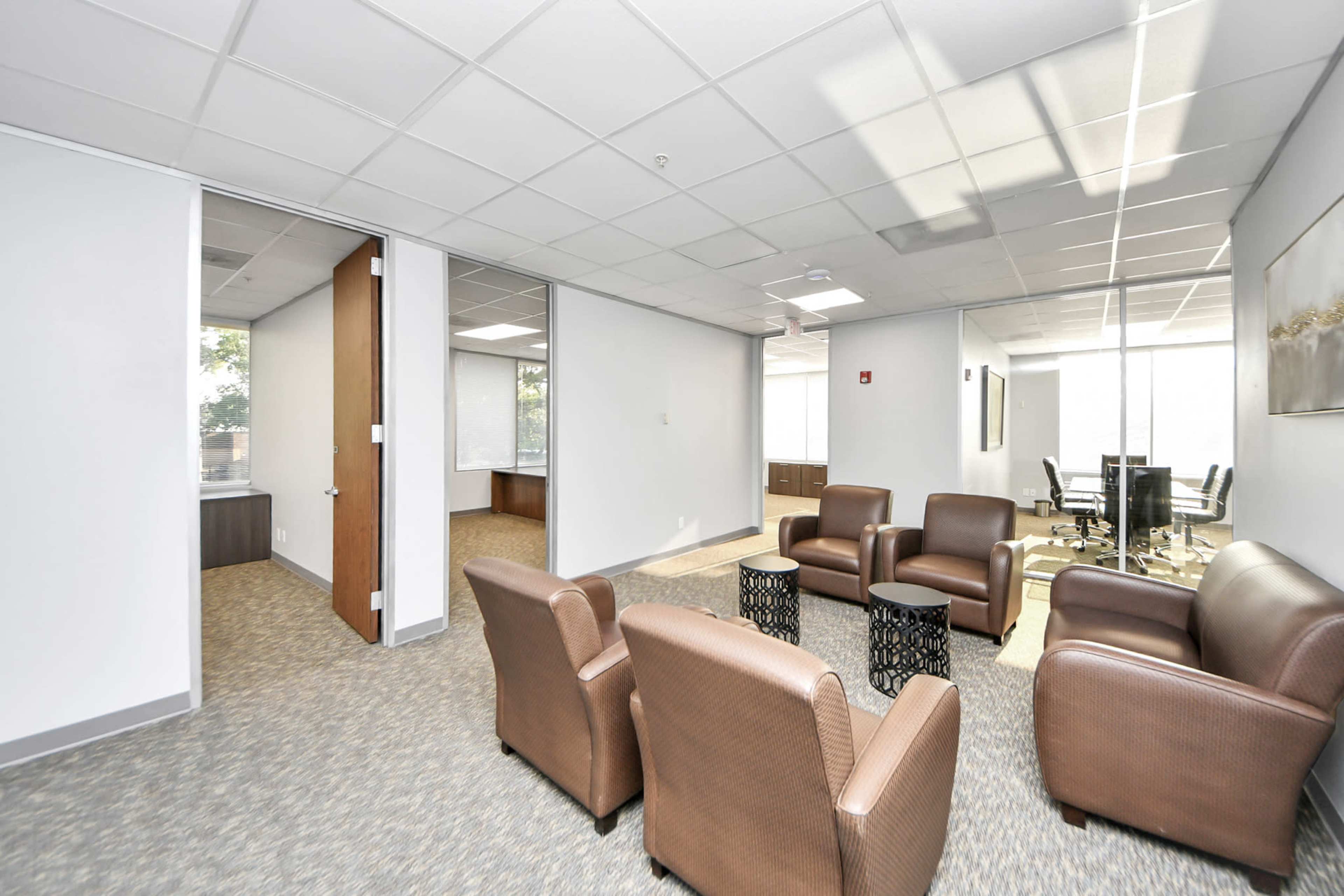 The image shows a modern office space featuring brown leather chairs arranged around a small table, with a conference room visible in the background.