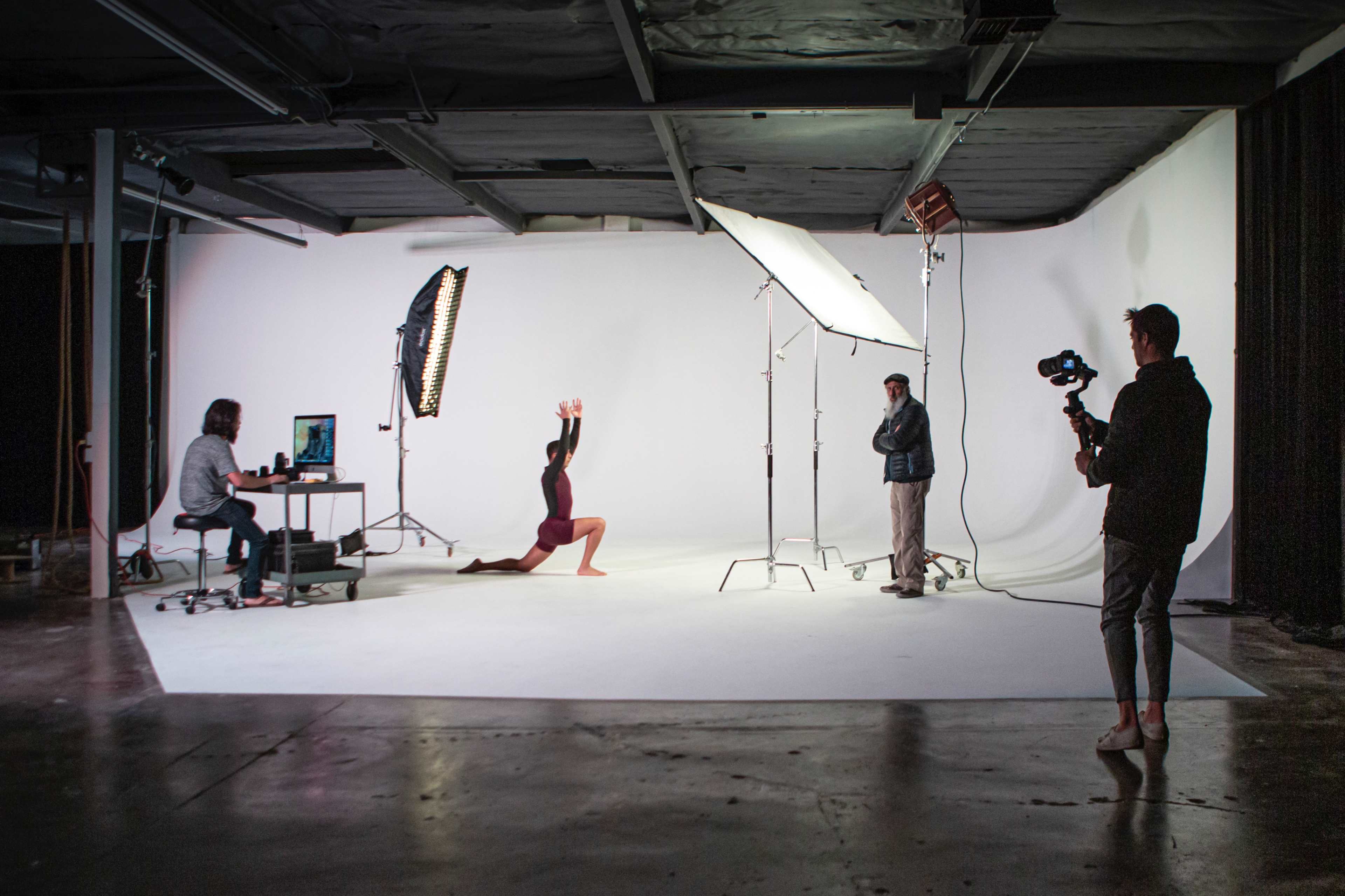 A photoshoot setup features a model posing on her knees in front of a white backdrop, while a photographer and an assistant adjust lighting equipment, and another person operates a camera.