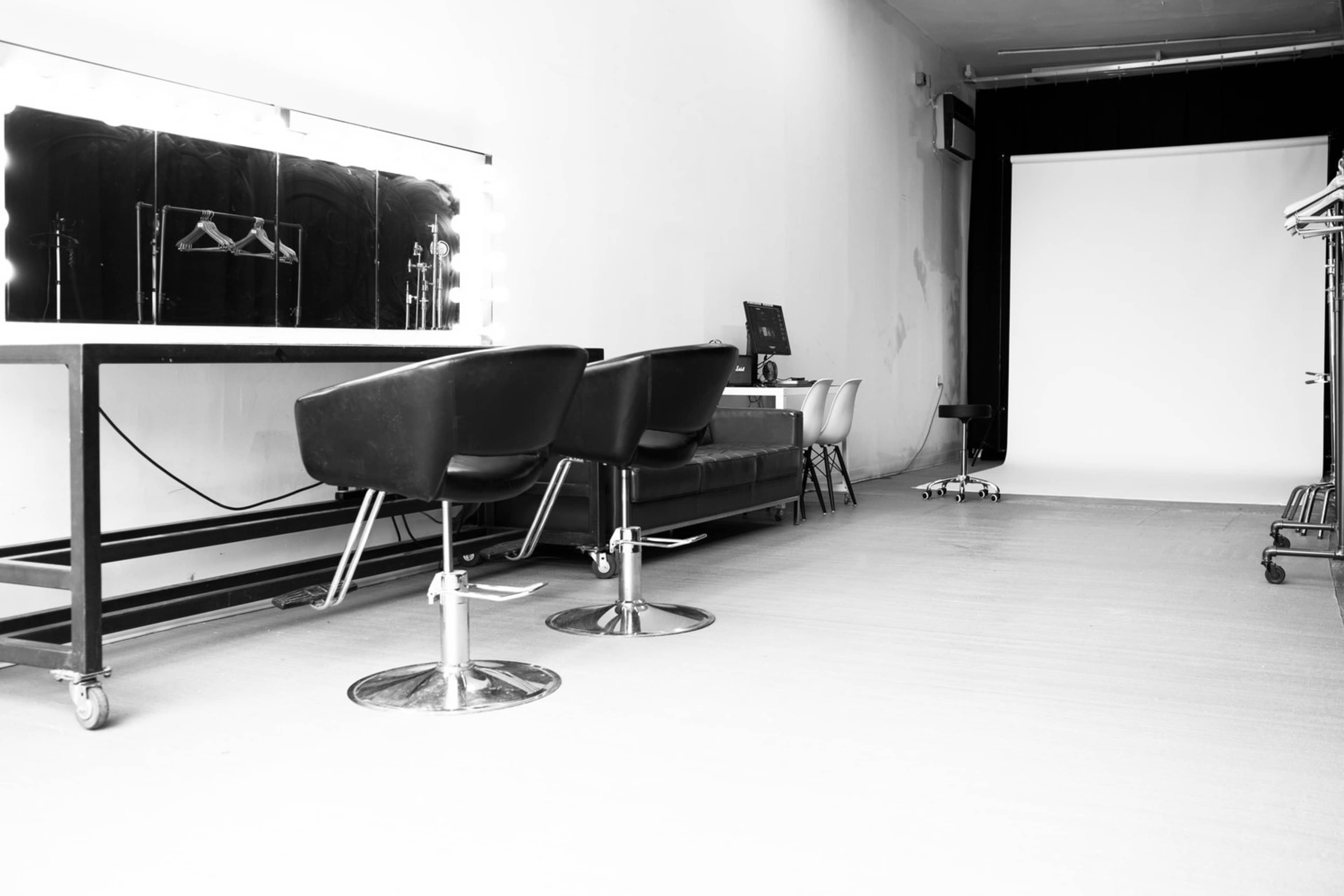 A minimalist studio space featuring a large illuminated mirror, two black salon chairs, and a backdrop.