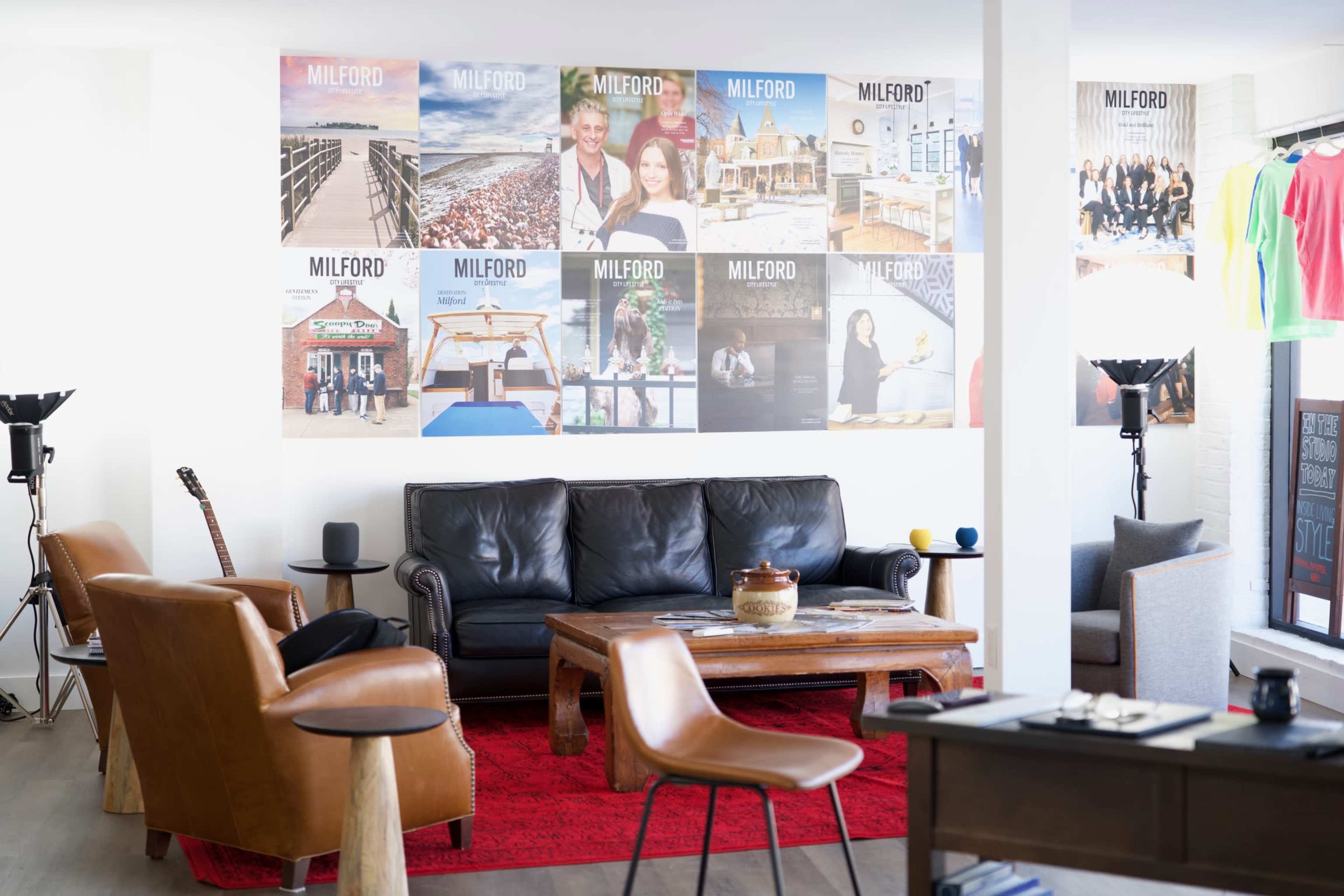 The image shows a modern seating area with a black leather sofa, a wooden coffee table, and several framed images on the wall, featuring various scenes and people.