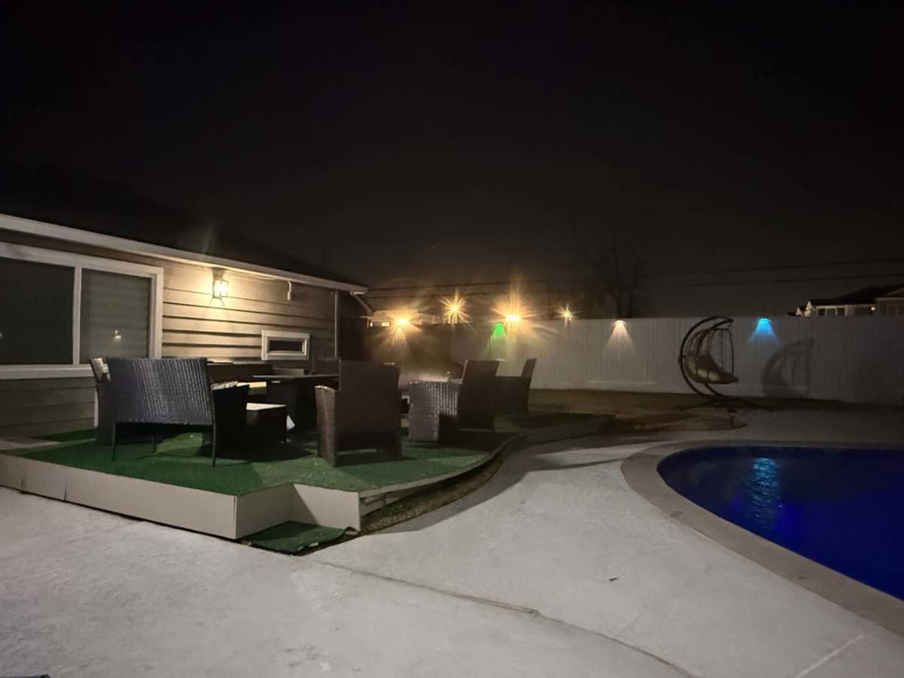 A patio area with wicker chairs and a small table is situated beside a pool, illuminated by decorative lights against a night sky.