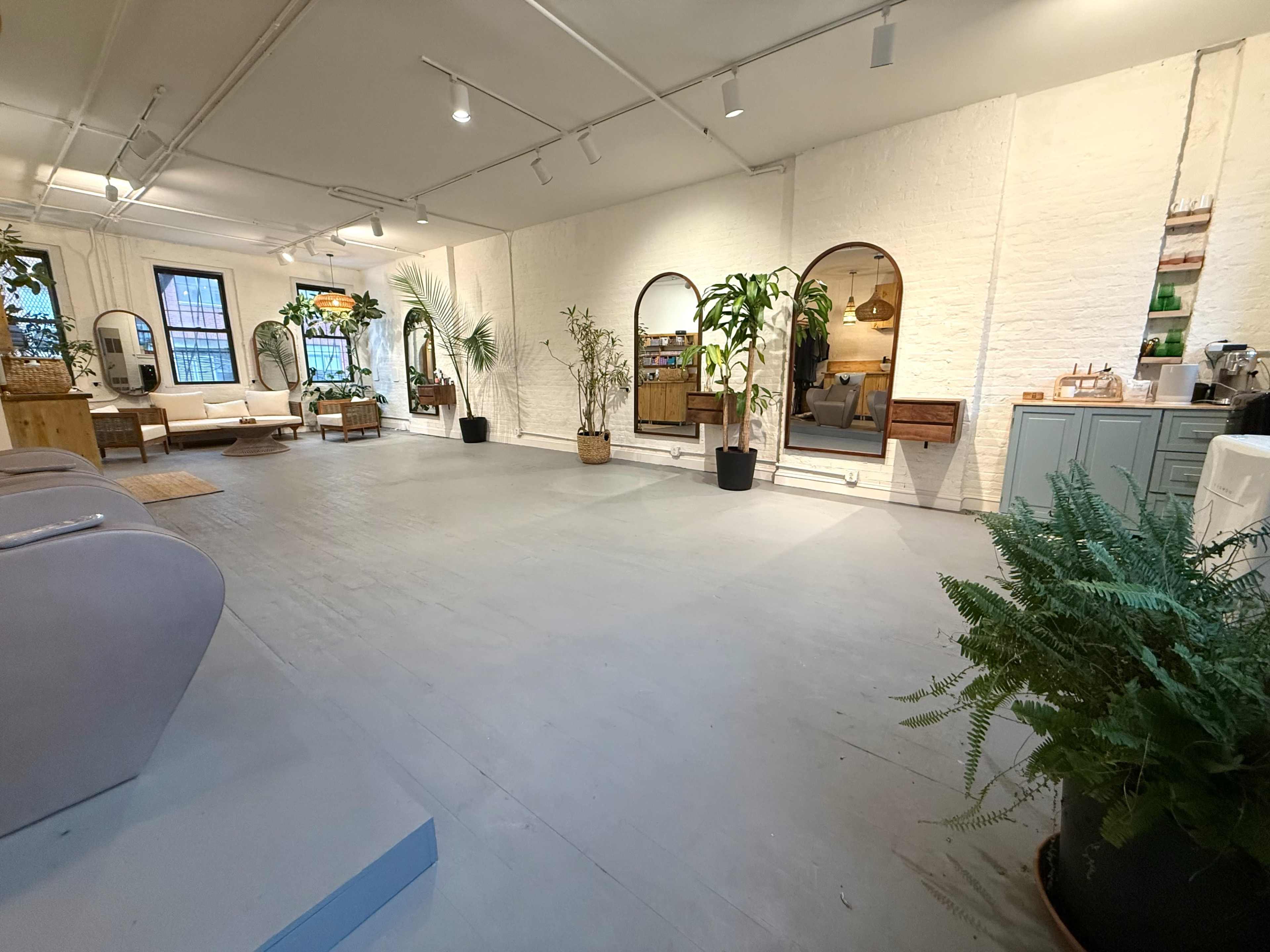 The image shows a spacious, well-lit indoor area featuring minimalist furniture, plants, and decorative mirrors against white brick walls.