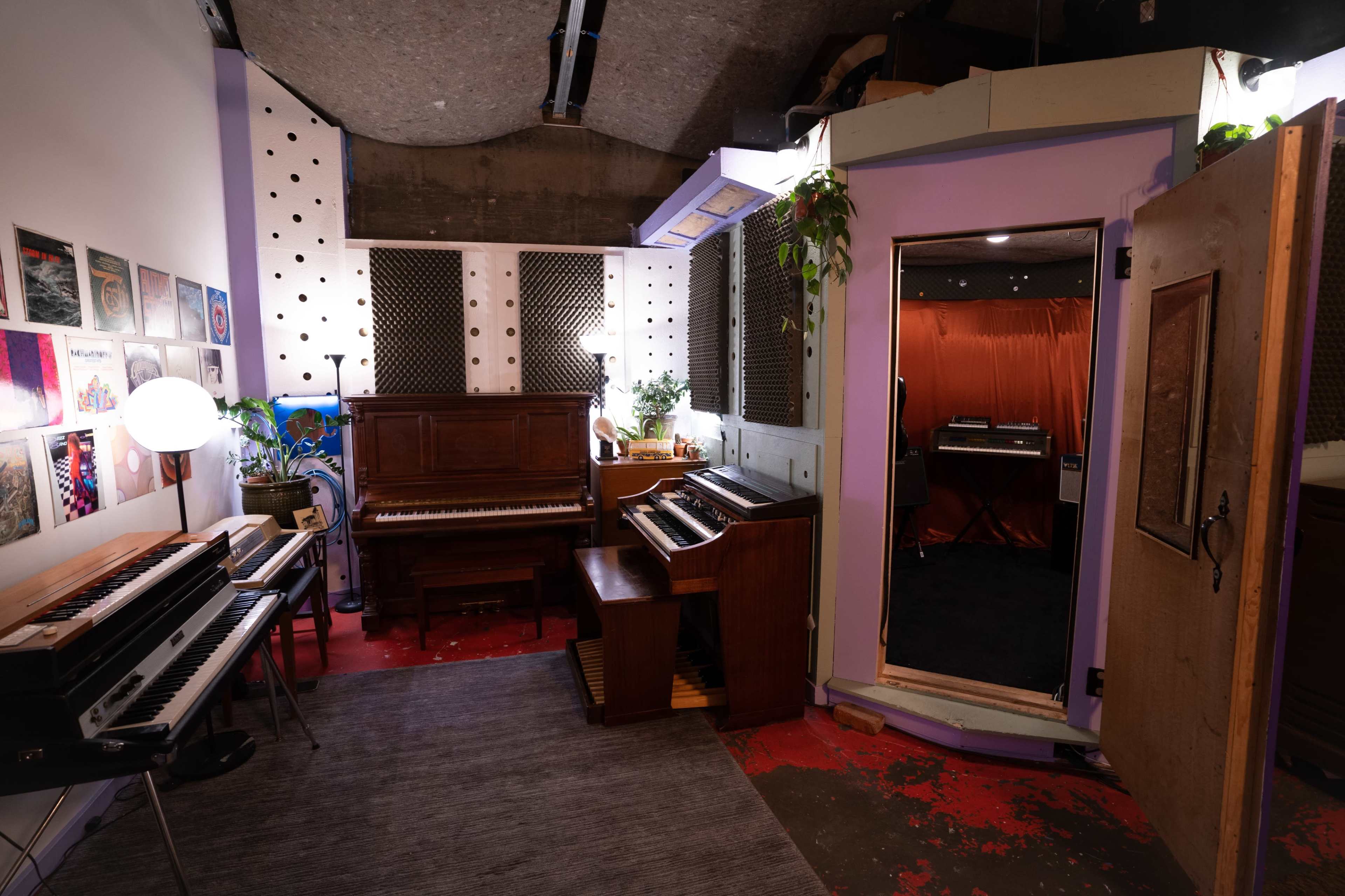 A music studio featuring multiple keyboards, a piano, and a booth for recording, with plants and wall art enhancing the space.