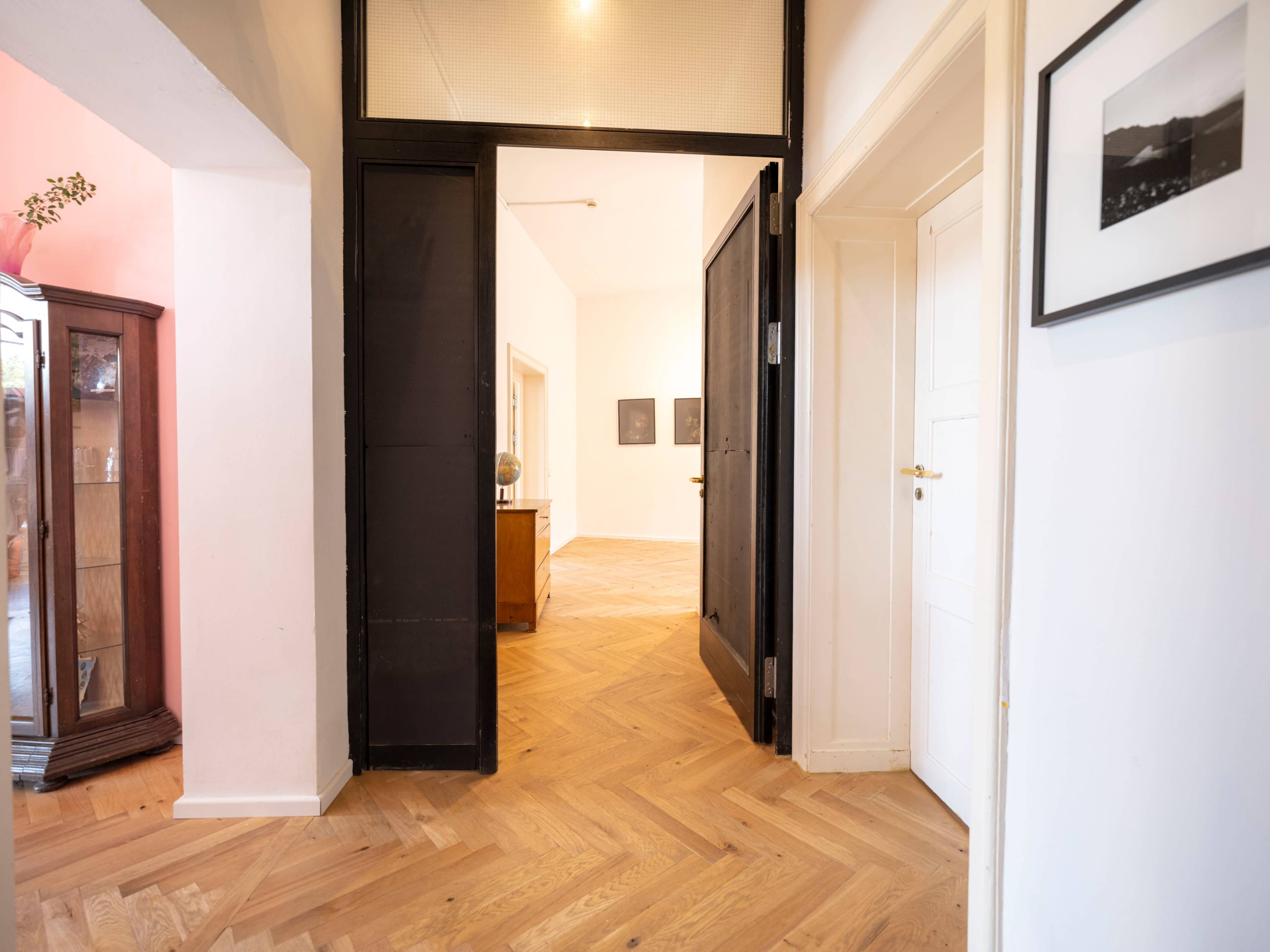 The image shows a spacious hallway with wooden herringbone flooring, leading to two open doorways on either side.