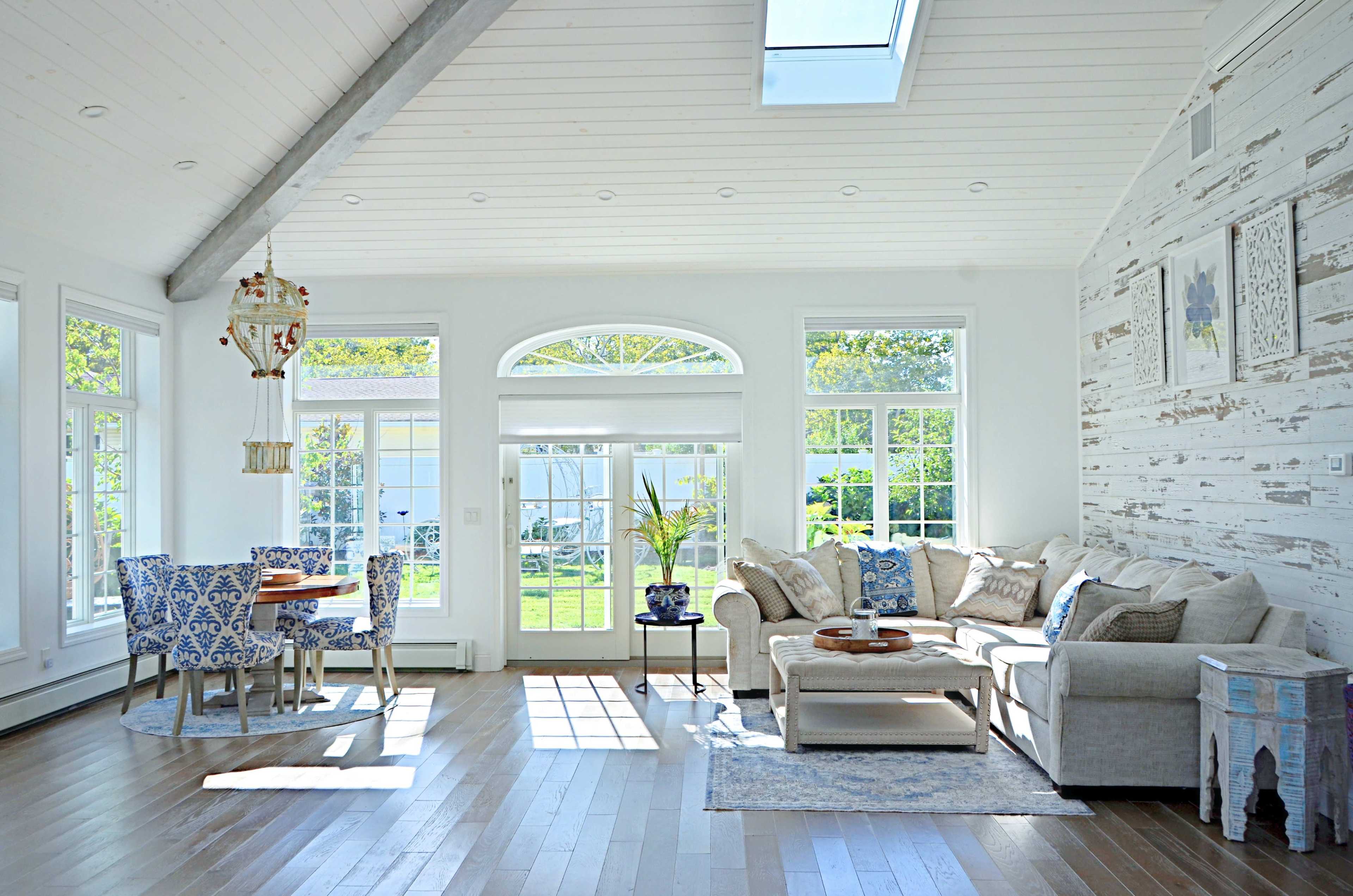 The room features a bright, open space with large windows, a sectional sofa, a wooden coffee table, and a dining area with blue patterned chairs.