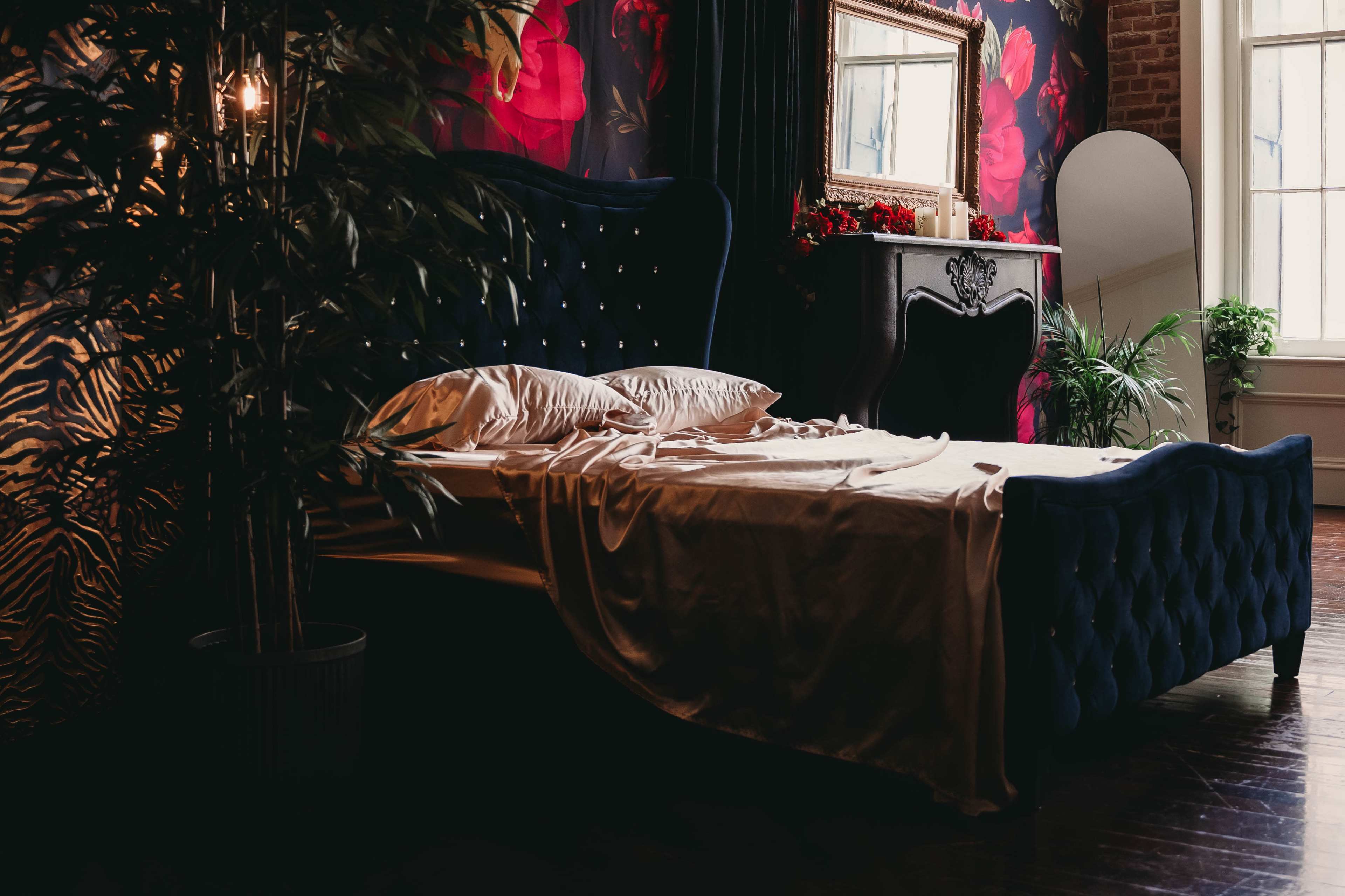 A dark blue upholstered bed sits against a floral wallpaper backdrop, featuring pillows and a crumpled satin blanket, with a vintage mirror and decorative plants nearby.