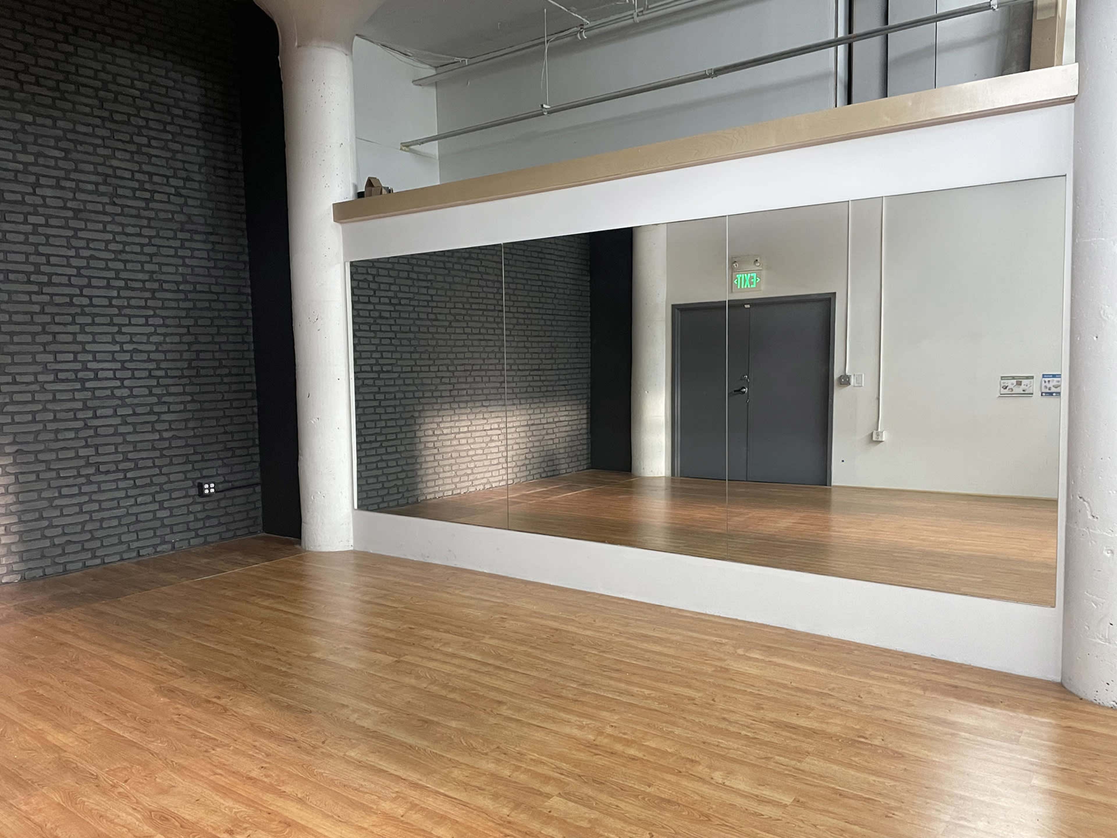 The image shows a spacious room with wooden flooring, a large mirror mounted on one wall, and a dark brick wall opposite it.