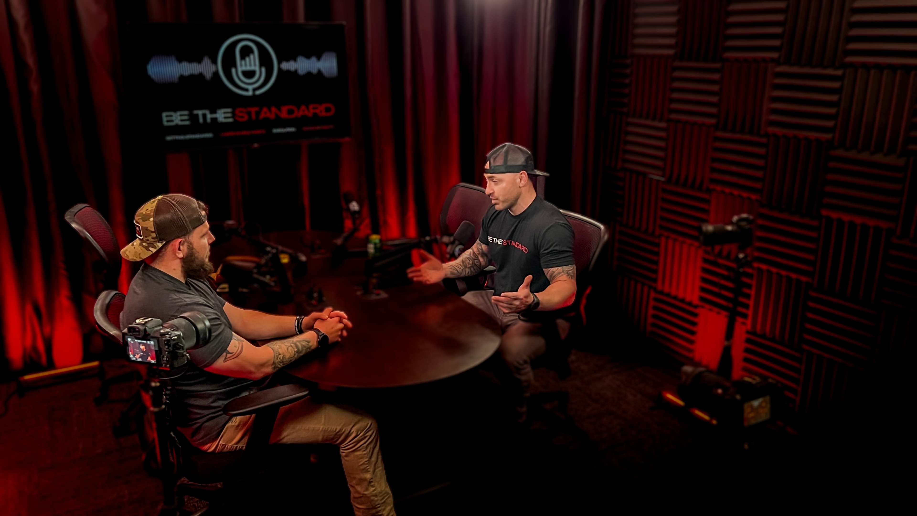 Two men are seated at a round table in a recording studio, discussing with microphones in front of them, while a screen behind them displays "BE THE STANDARD."