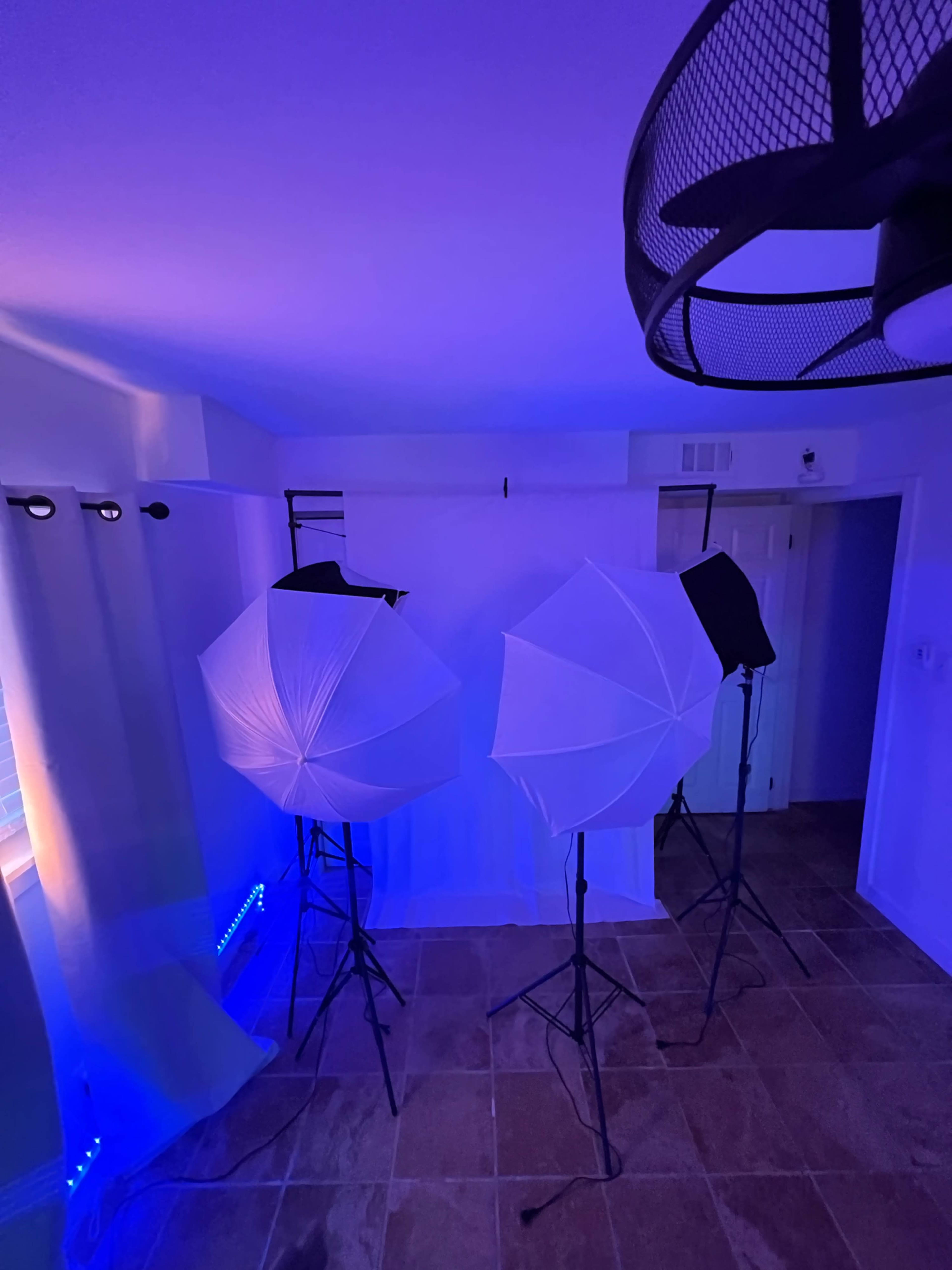 The scene features two light umbrellas on stands facing a backdrop in a room with blue LED lighting and a ceiling fan.