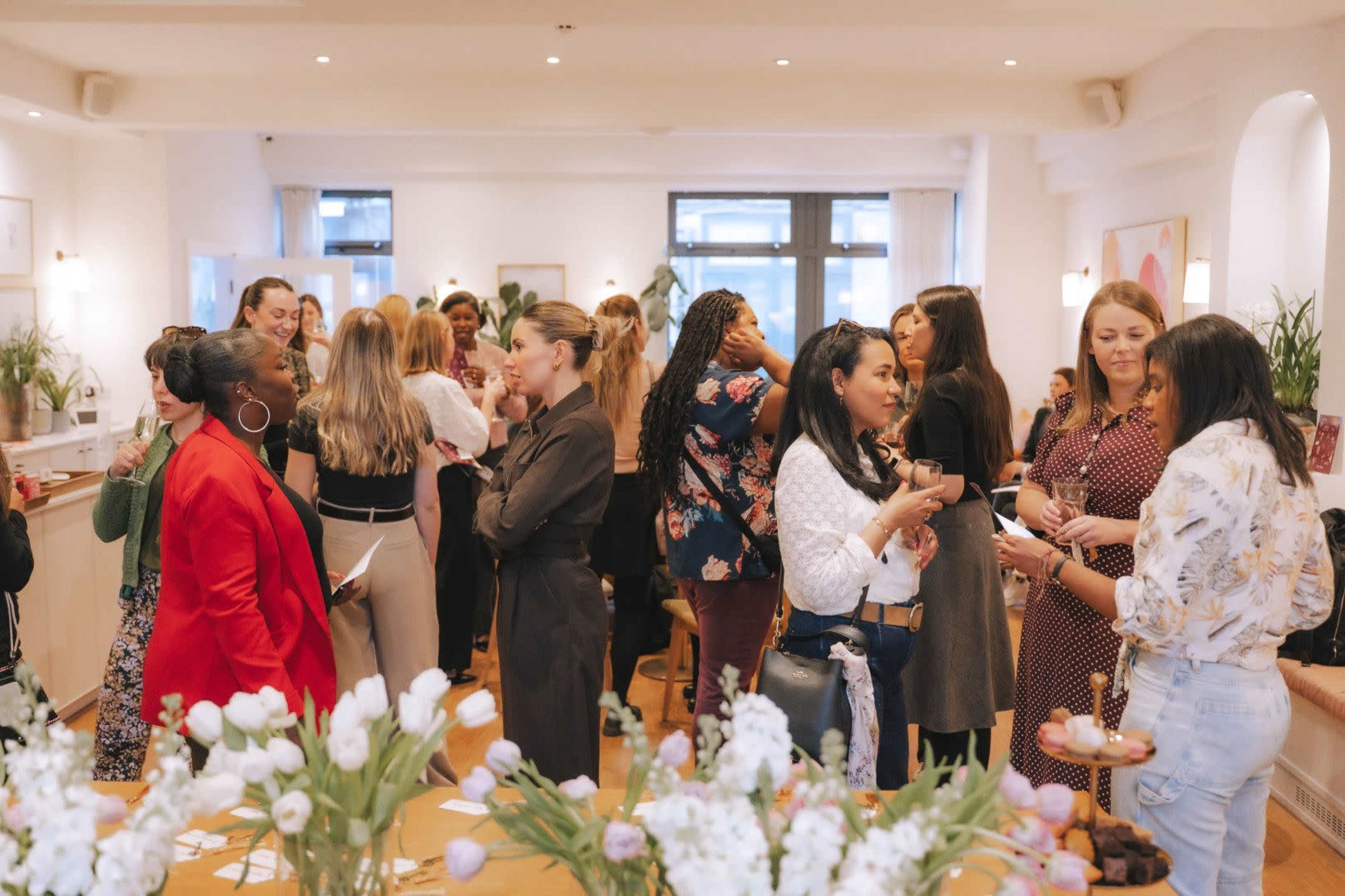 A group of women is mingling and socializing at a brightly lit indoor event, with tables adorned with flowers and refreshments.