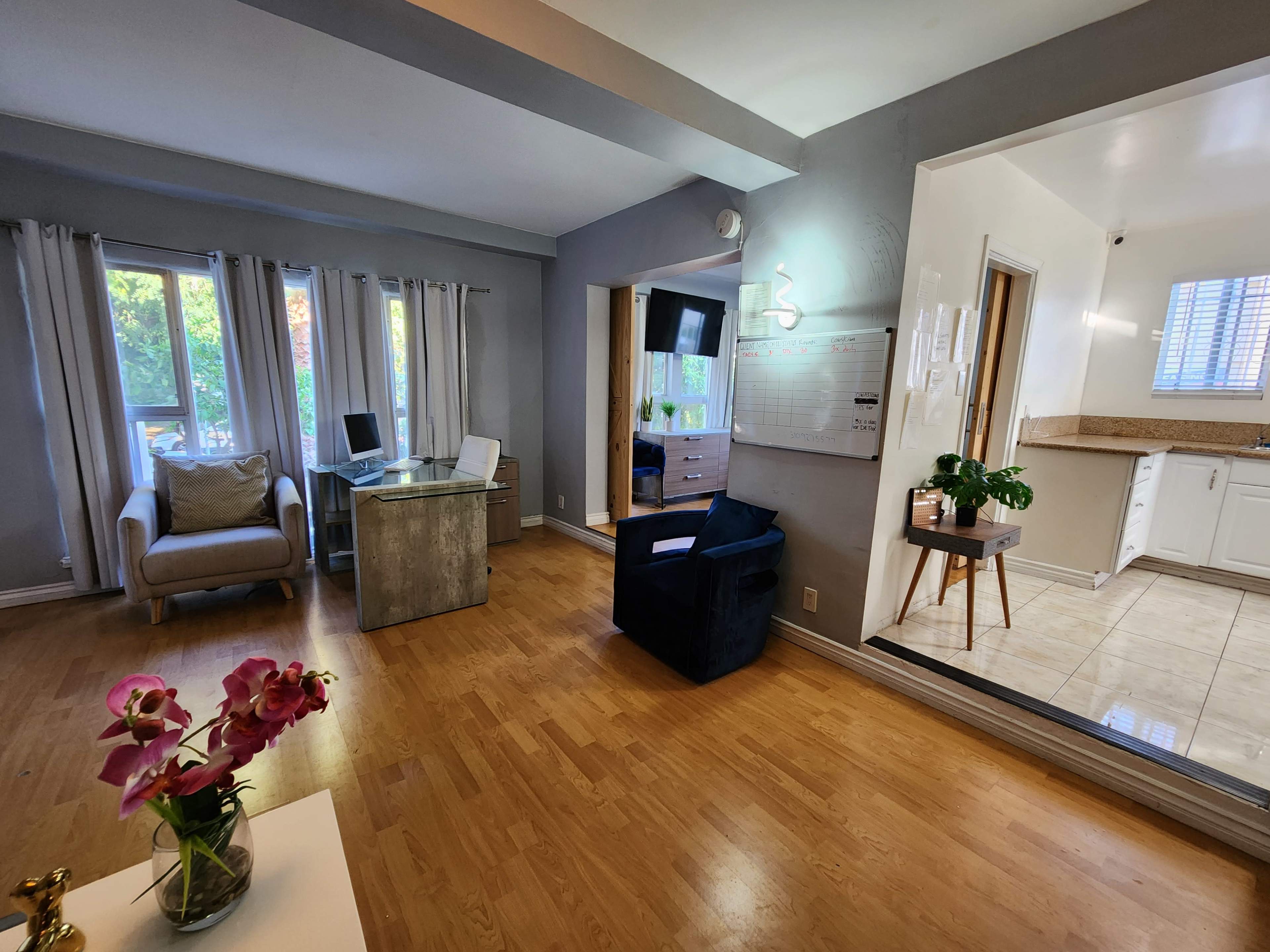 The image shows a modern living space featuring a desk with a computer, a comfortable chair, and a well-lit kitchen area visible in the background.