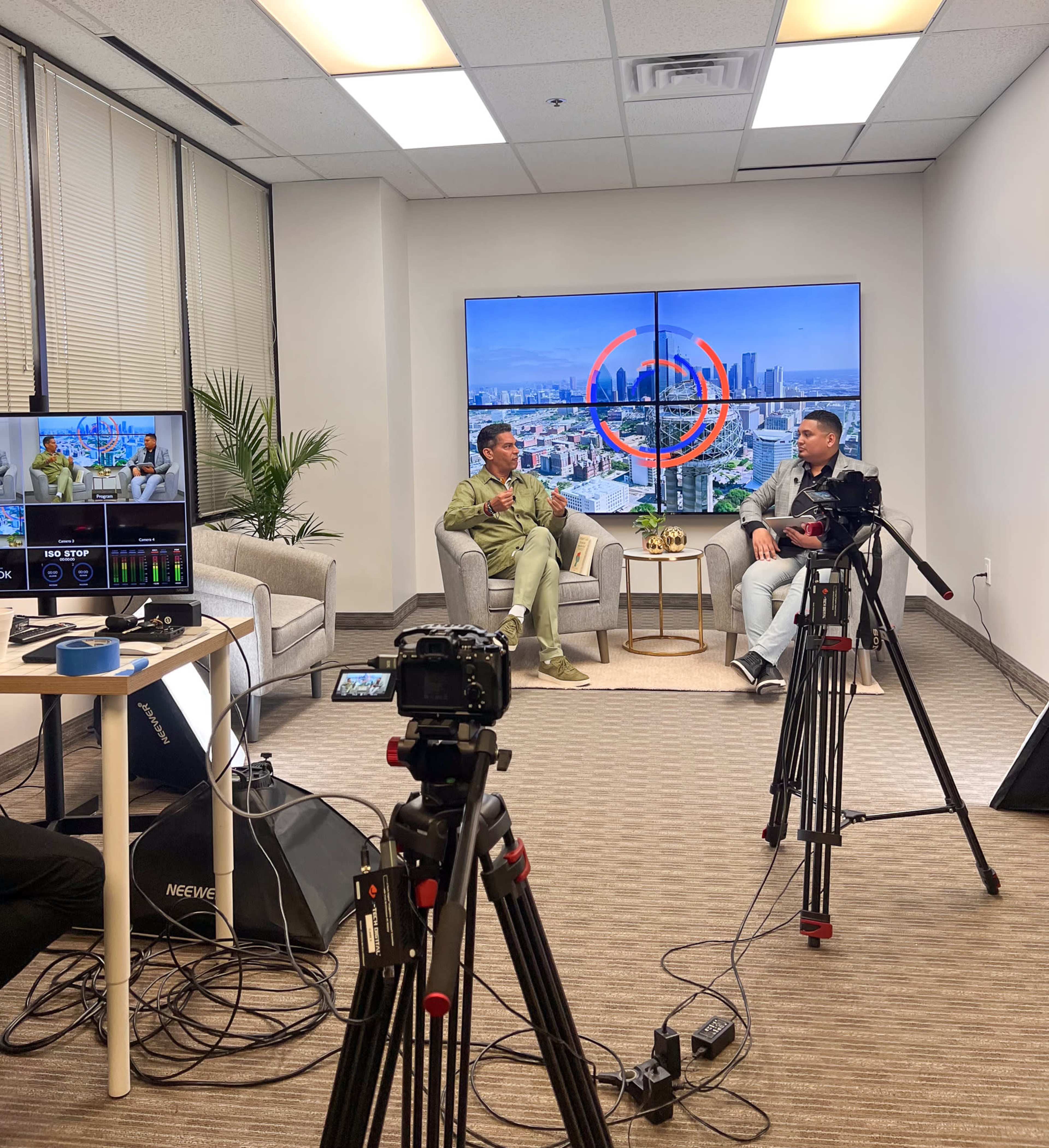 Two men are engaged in a conversation on a set with two large screens displaying a cityscape in the background, while cameras and equipment are set up around them.