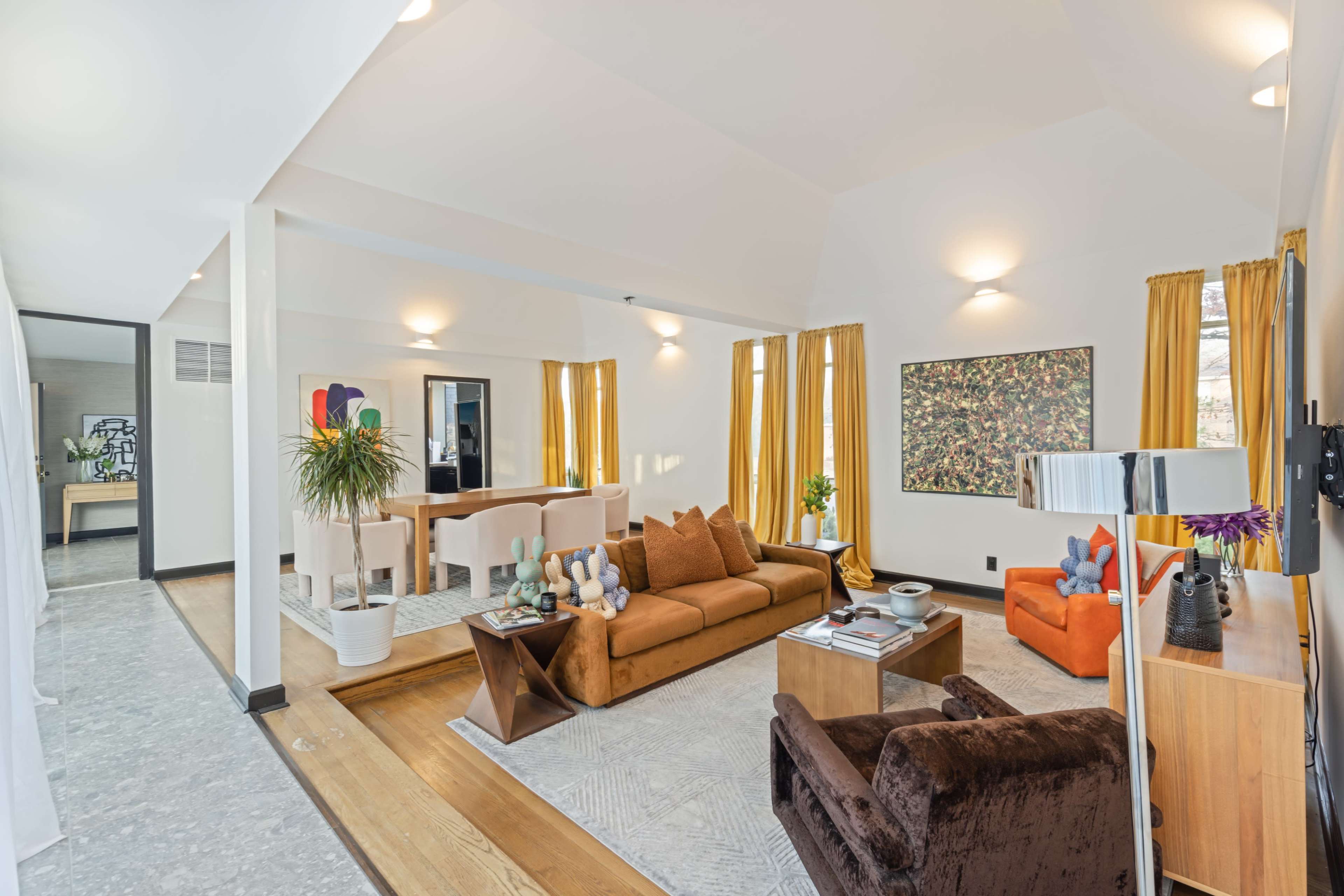 A modern open-concept living space featuring a brown sofa, an orange armchair, a dining area with a light table, and large windows with yellow curtains.
