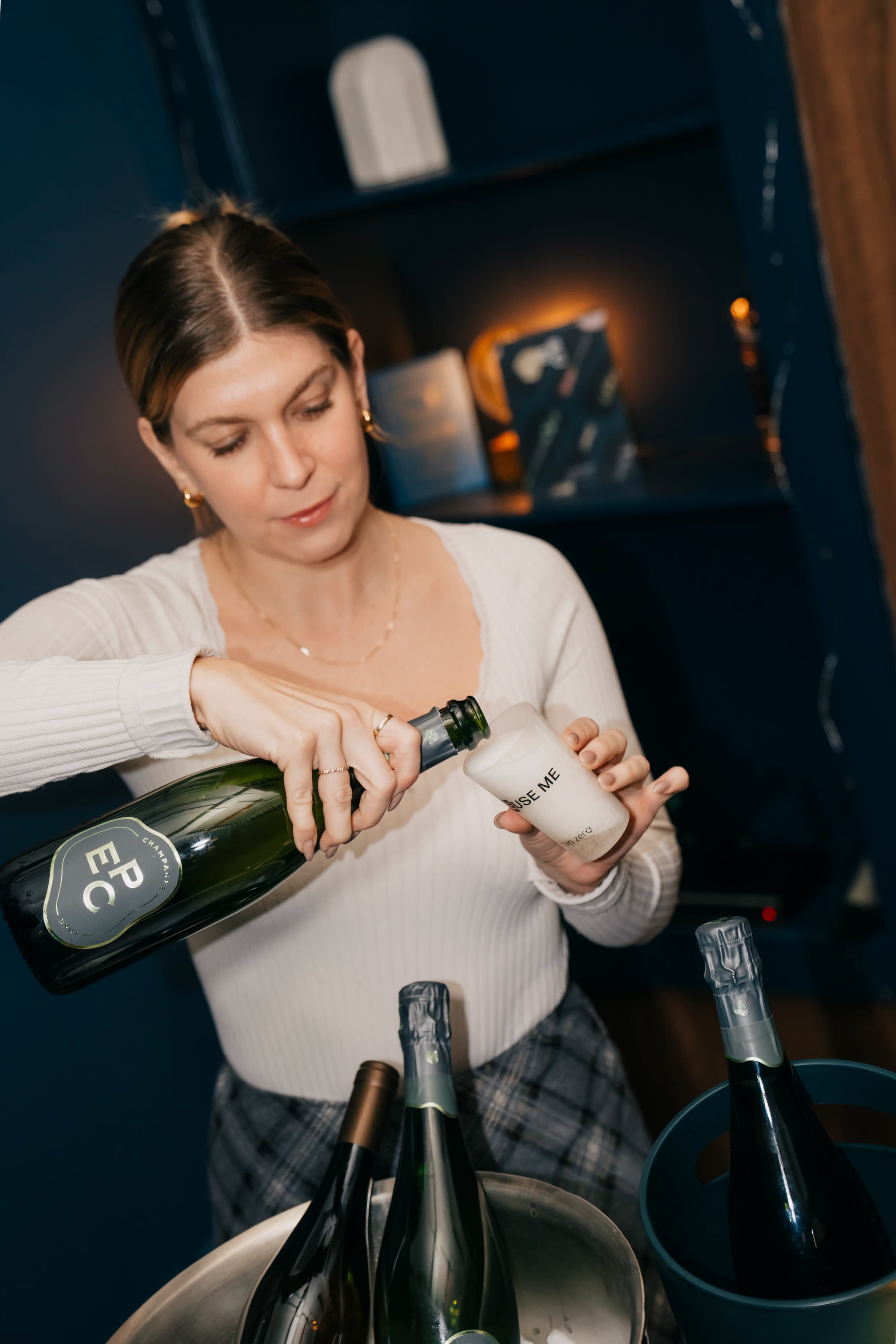 A woman in a white sweater pours sparkling wine from a bottle into a glass in a dimly lit room.