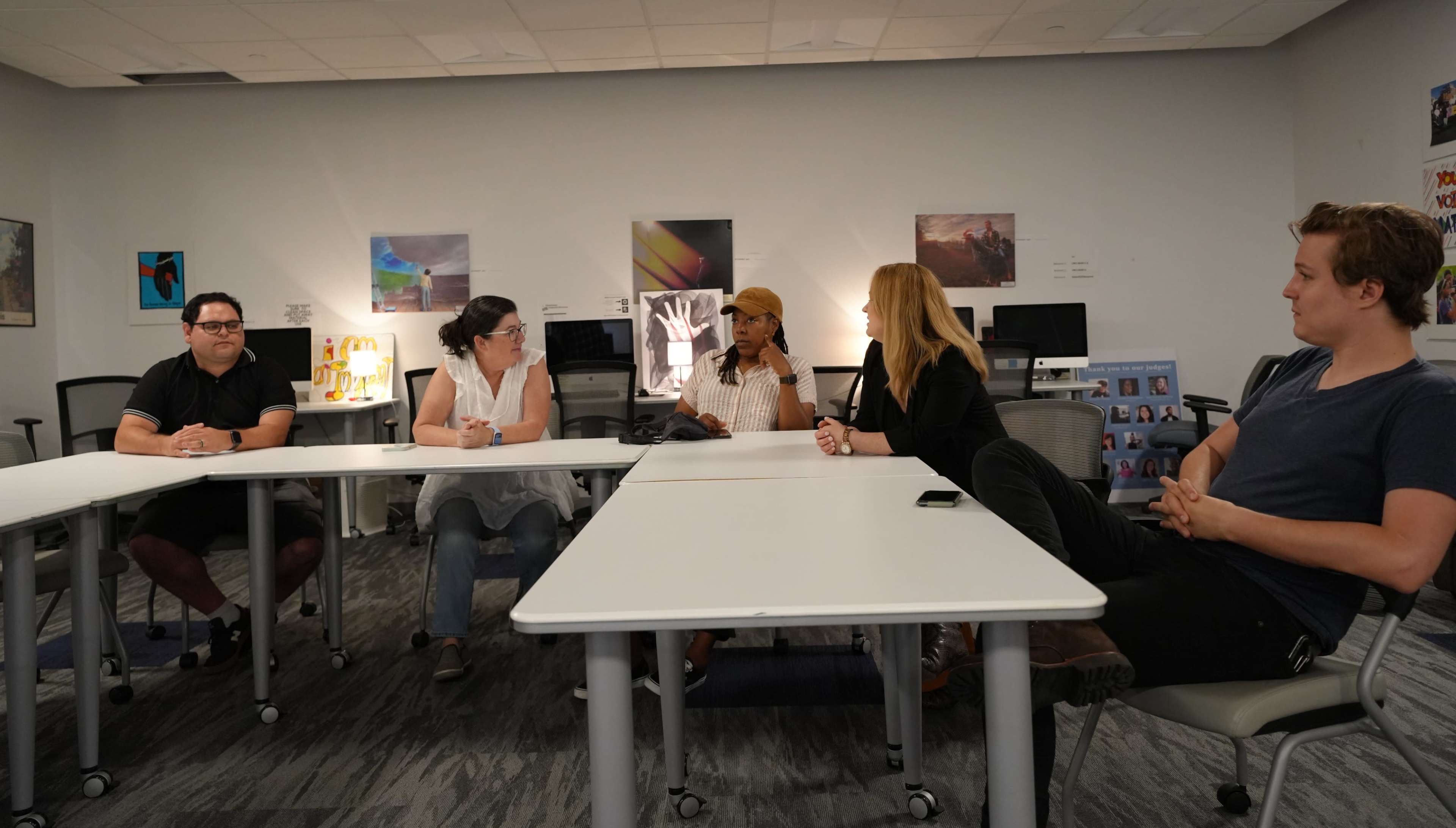 A group of five people sit around a table in a classroom setting, engaged in discussion with various artworks displayed on the walls.