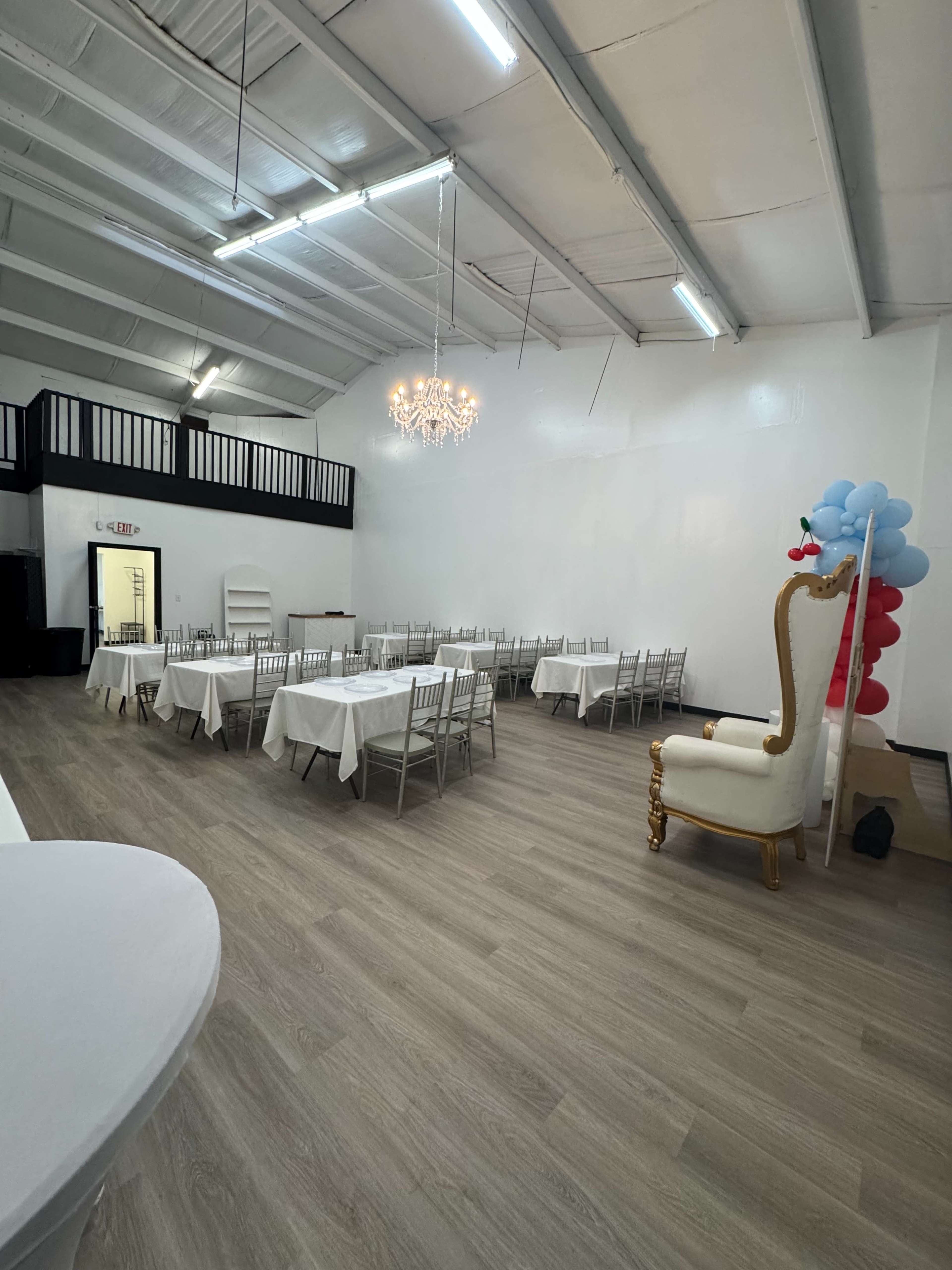 The image shows a spacious event room with white walls, a chandelier, and rows of tables set with white tablecloths, accompanied by a large decorative chair and balloon decorations.