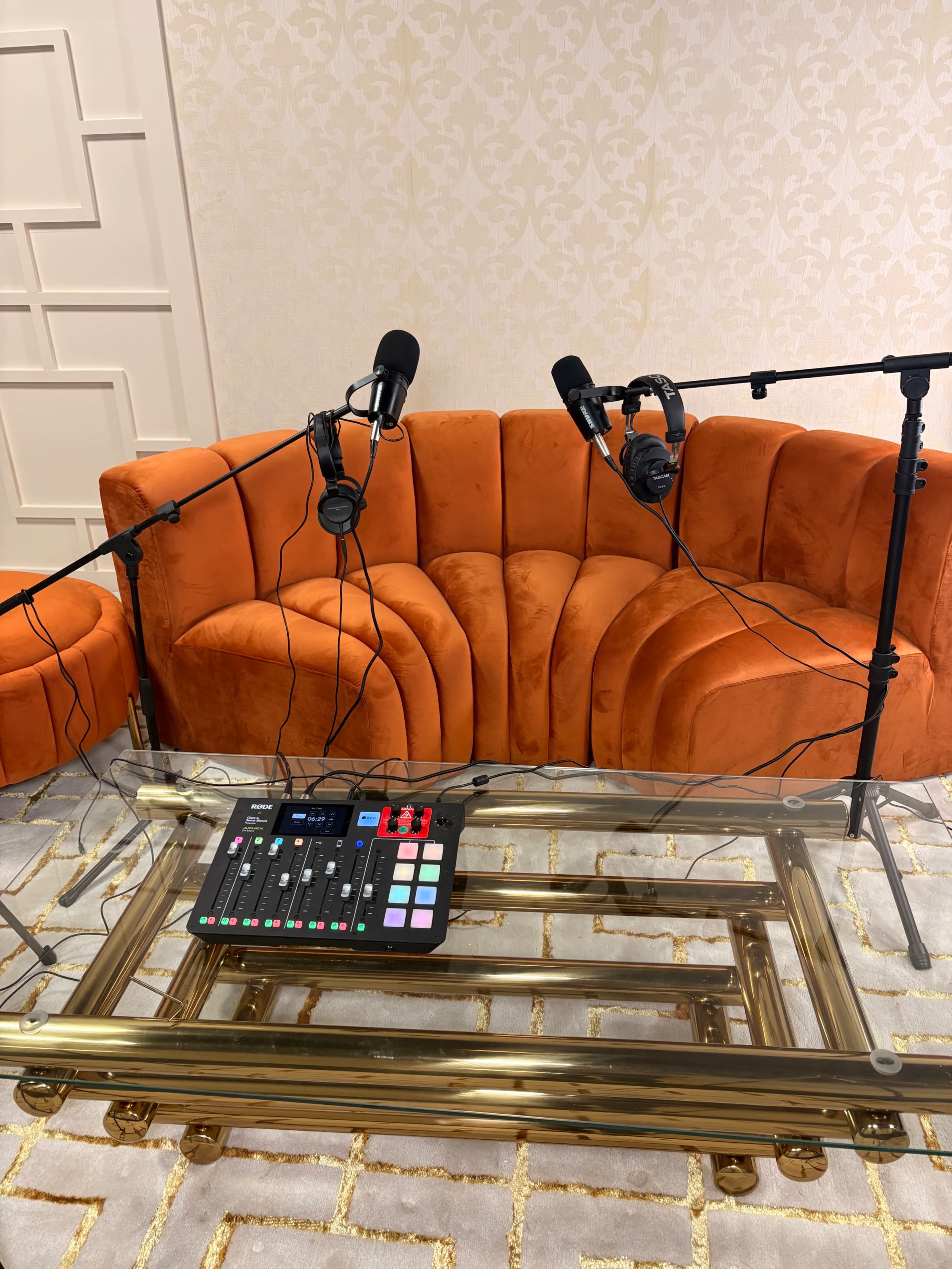 A vibrant orange couch with two microphones mounted on stands and a sound mixing board on a glass coffee table is set up in a well-lit room.
