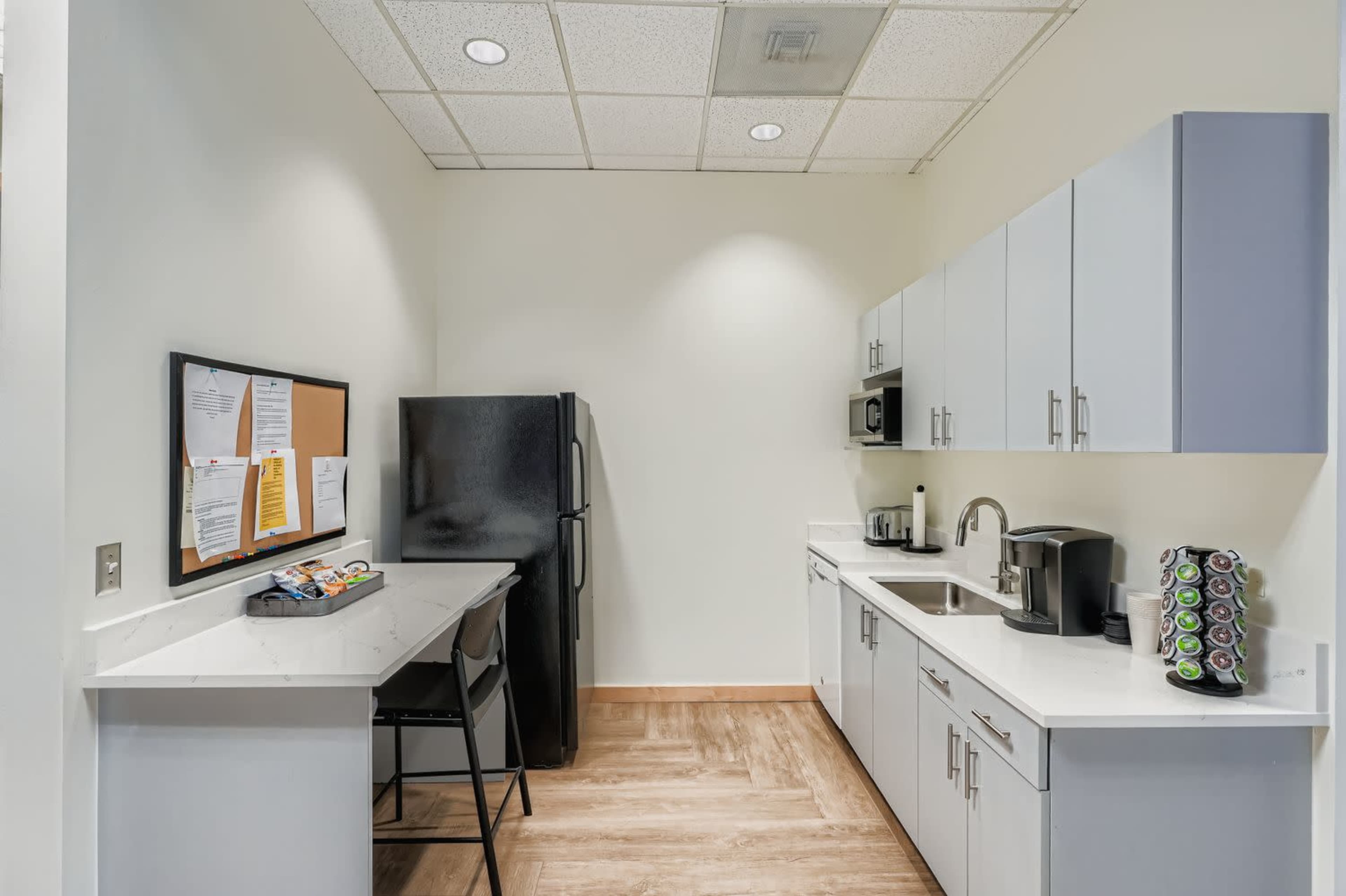 The image shows a small kitchen area with a black refrigerator, a sink, a microwave, and a coffee maker, along with a counter and a seating area.