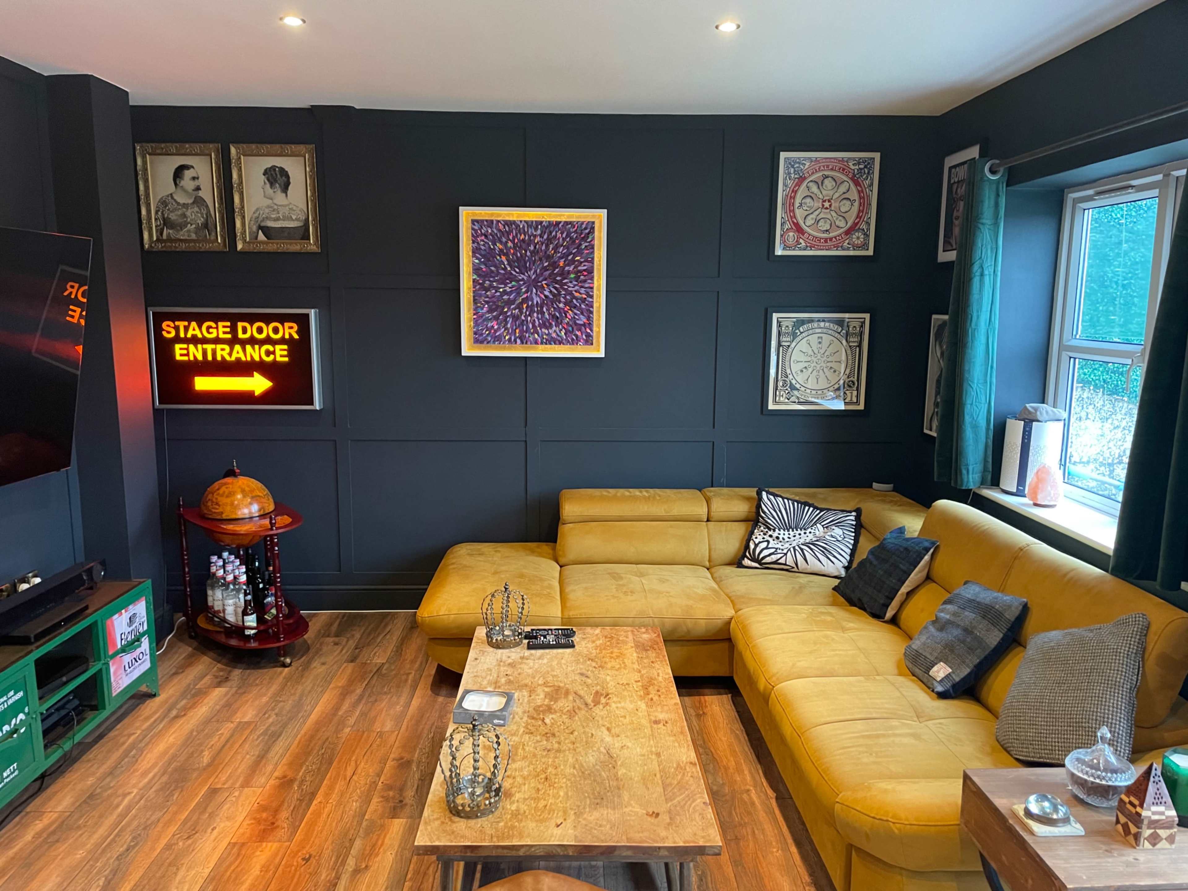 The image shows a cozy living room featuring a yellow sectional sofa, a wooden coffee table, and various framed artworks on the dark wall, along with a glowing sign that reads "STAGE DOOR ENTRANCE."