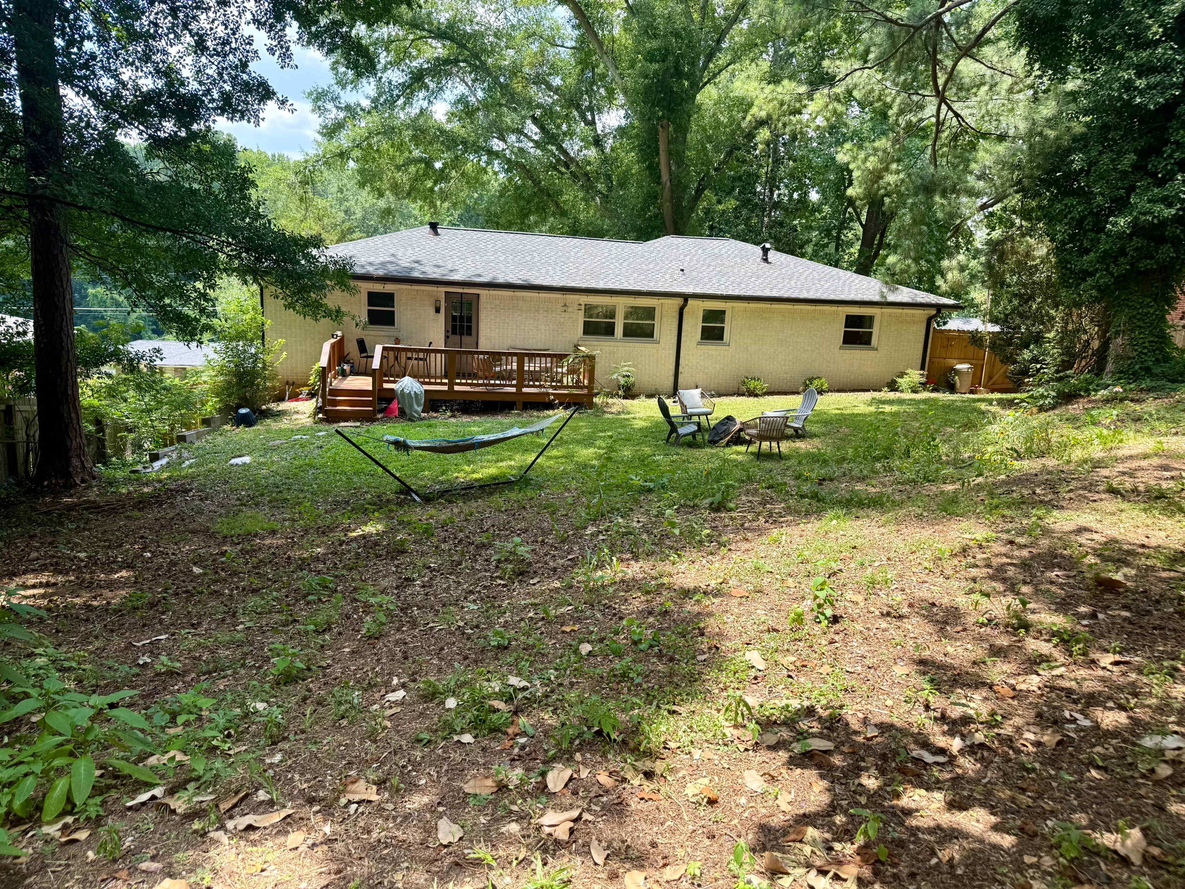 A backyard features a house with a deck, surrounded by trees, with a hammock and several lawn chairs visible on the grassy area.