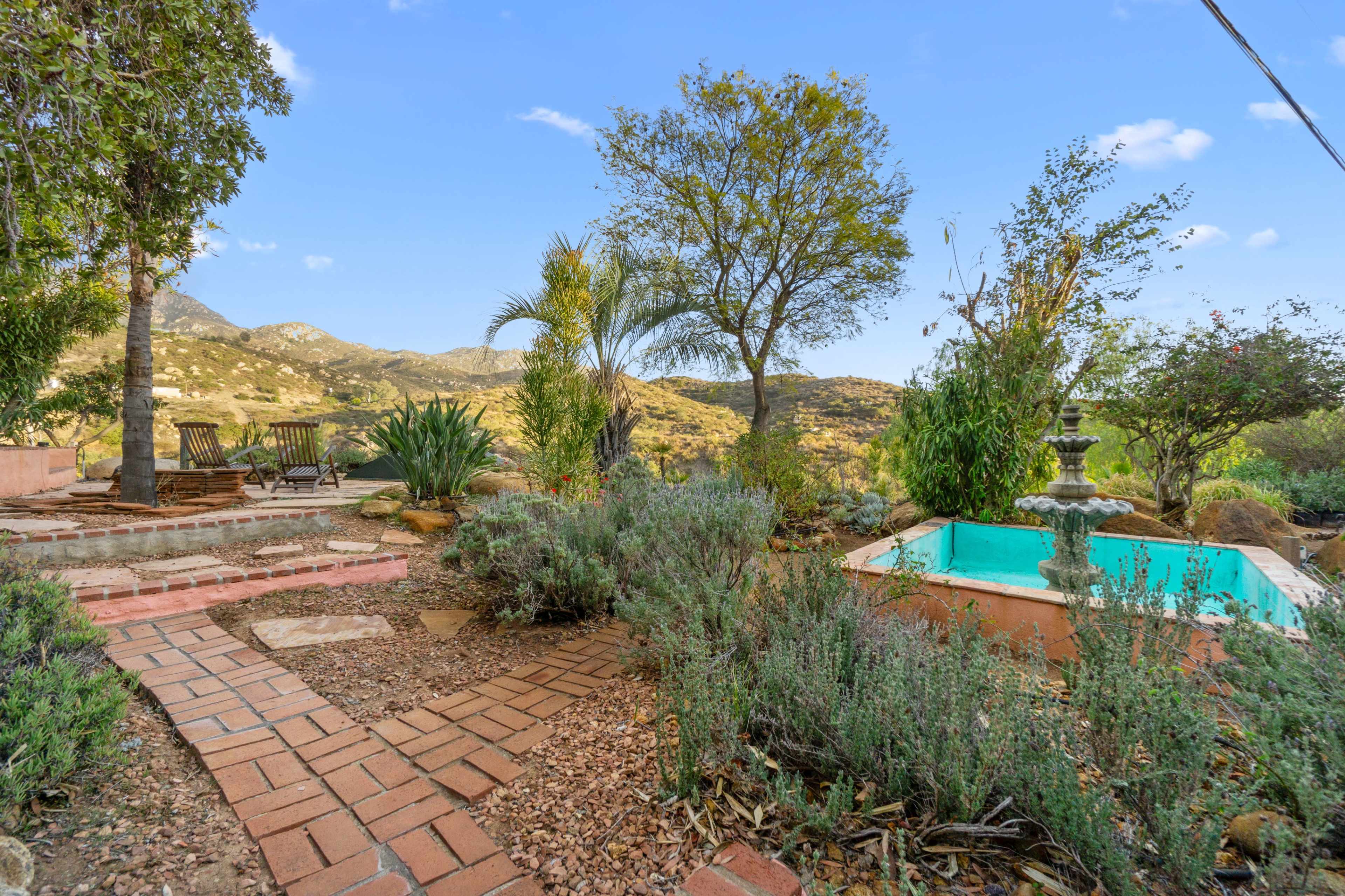 The image shows a landscaped outdoor garden area with a brick pathway, various plants, trees, and a small turquoise pool or fountain surrounded by natural scenery.