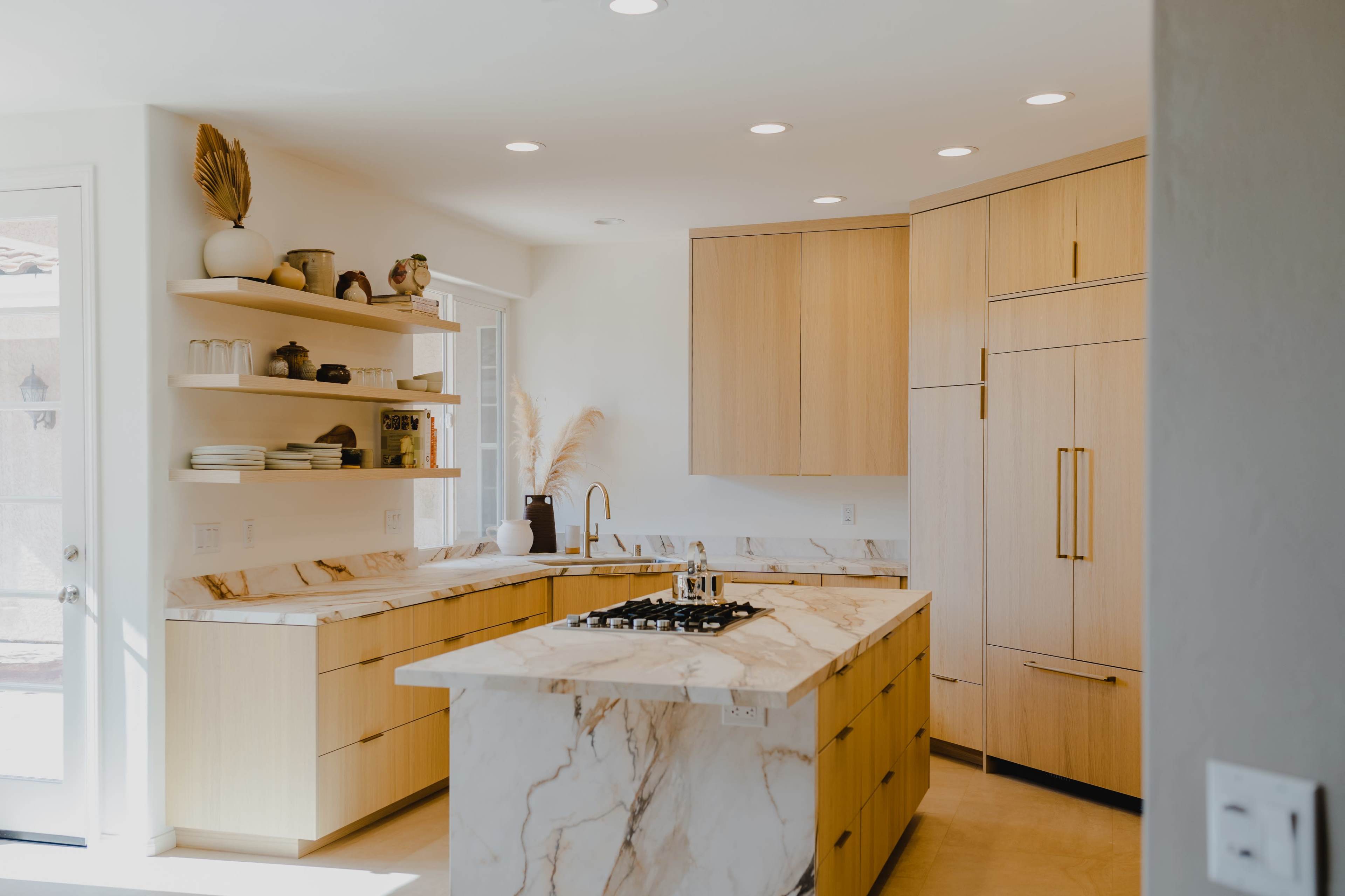 The kitchen features a marble island with a built-in gas cooktop, wooden cabinetry, and open shelving displaying various dishes and decor.