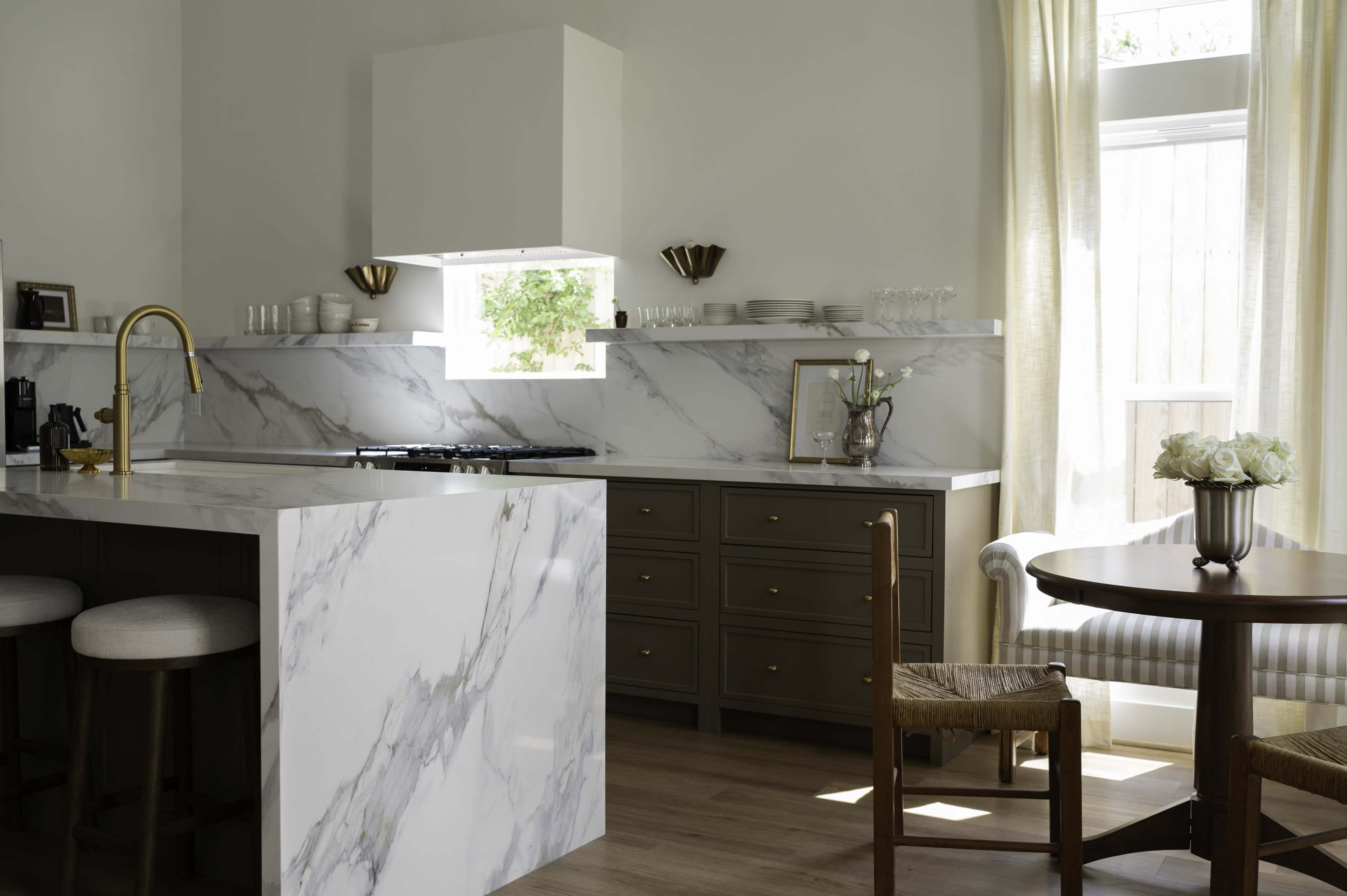 The image shows a modern kitchen featuring white marble countertops, wooden cabinets, and a dining area with a round table and a soft sofa.